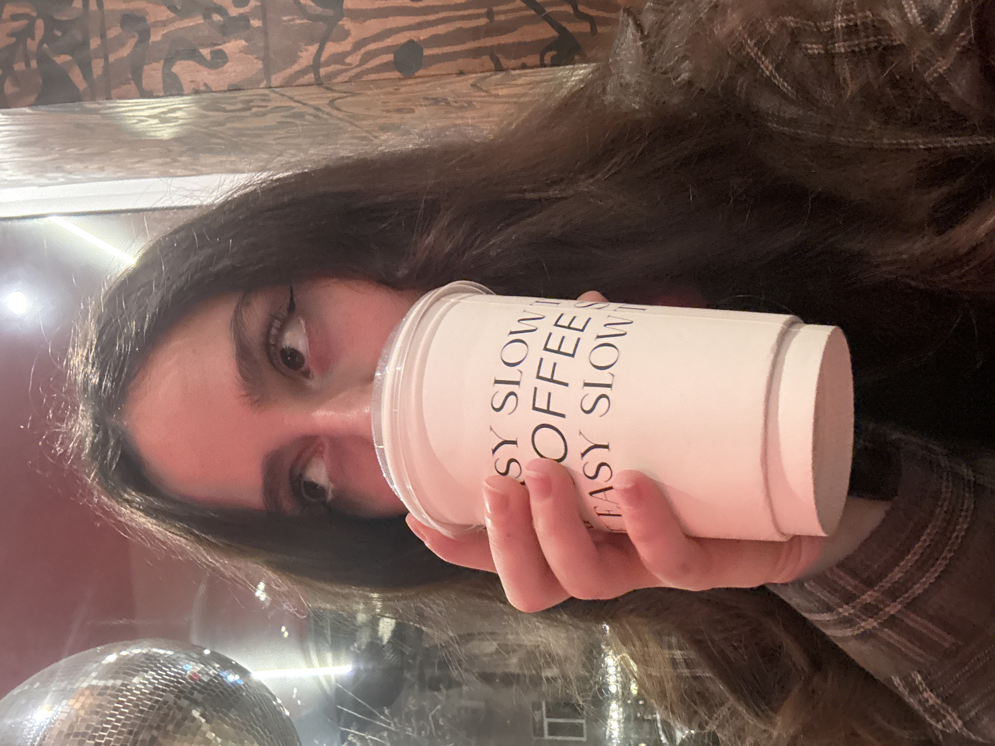 An individual, visible from the upper chest to the top of the head, is depicted indoors at Easy Slow Coffee in Yerevan, Armenia. The subject, a person with long, dark brown, wavy hair, holds a white disposable coffee cup to their mouth with both hands, appearing to drink. The cup bears black text, partially legible as "SY SLOW COFFEE EASY SLOW". The individual wears a brown and beige plaid shirt and looks upwards and to the right. To the left of the subject, a reflective surface, possibly a window, shows a blurry exterior scene with lights, and a spherical disco ball hangs prominently in the upper left quadrant. To the right, a wall with a dark, abstract pattern on a lighter brown background is visible. The scene is dimly lit with ambient light and reflections.