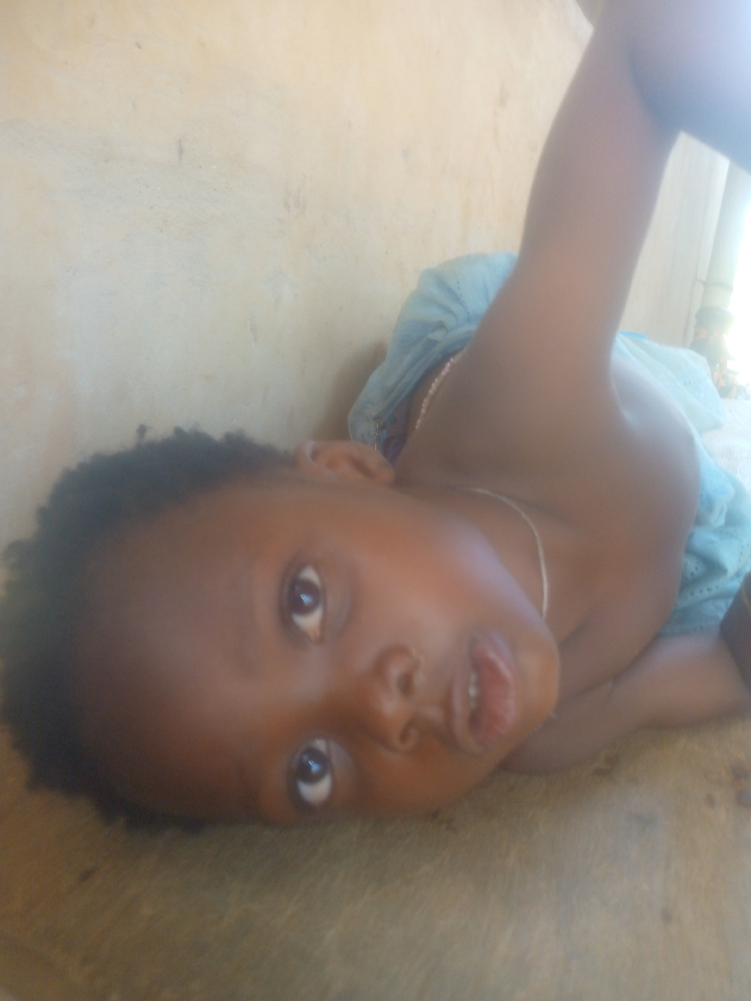 A young child with dark skin and short, dark hair is lying down, looking at the camera. The child is wearing a light blue top and a silver necklace. The background appears to be a plain, light-colored wall. The location is Unknown, Unknown.