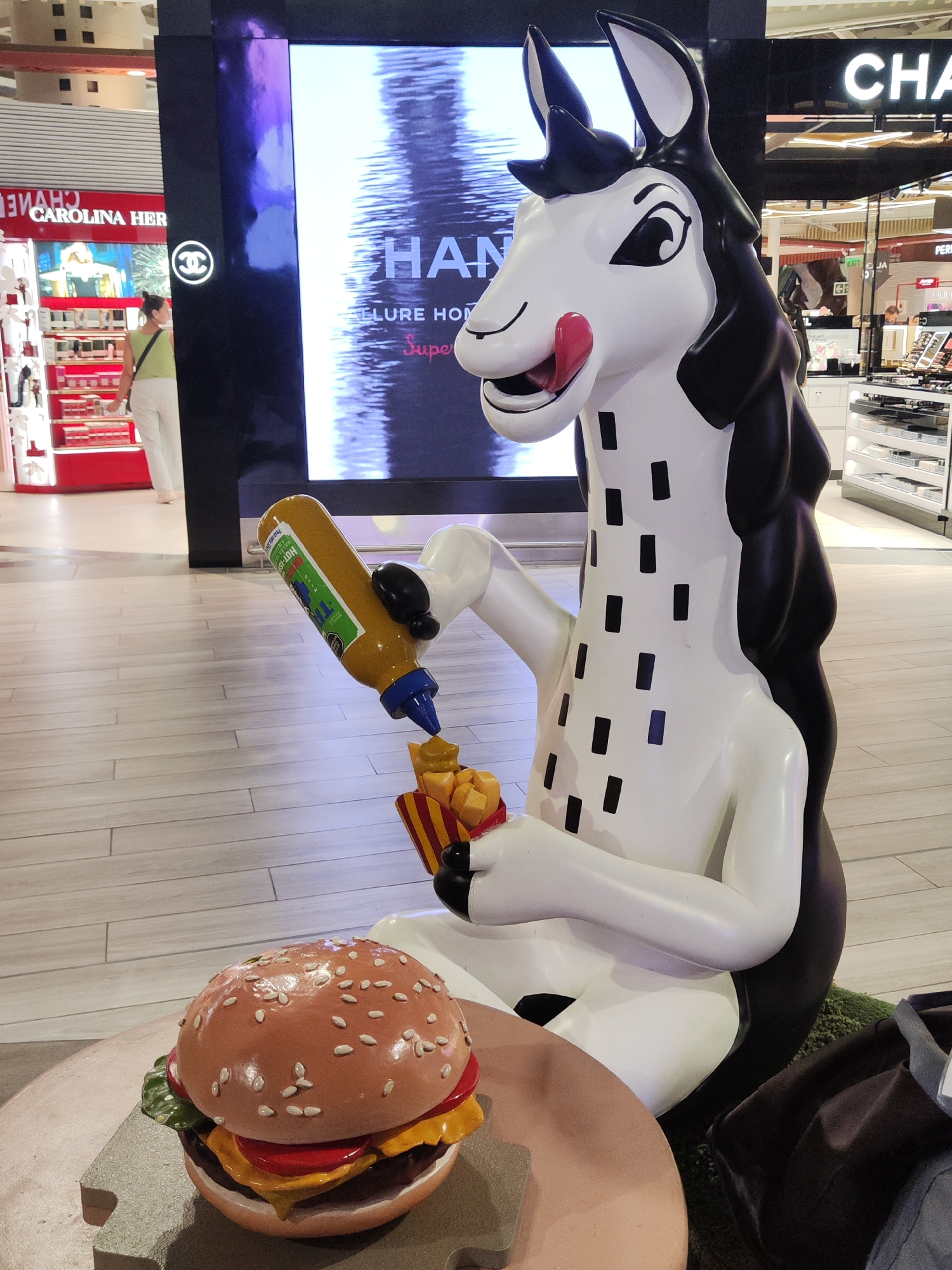 A full shot shows a cartoonish sculpture of a black-and-white llama. The llama is in a seated position, its body facing slightly to the right. Its head is white with black ears and mane, and its tongue is sticking out, as if licking its lips. The neck and body of the llama are white with evenly spaced vertical black stripes. The llama is holding a container of French fries in its left hand and a bottle of mustard in its right hand, and appears to be squeezing mustard on the French fries.
To the left of the llama is a large burger on a small round table.
The sculpture sits inside a shopping mall or similar retail environment. The sculpture is surrounded by retail store displays, including CAROLINA HER, CHANEL, and another store with the words "PER" and "CHA" visible. Other shopping mall patrons are also present in the background.