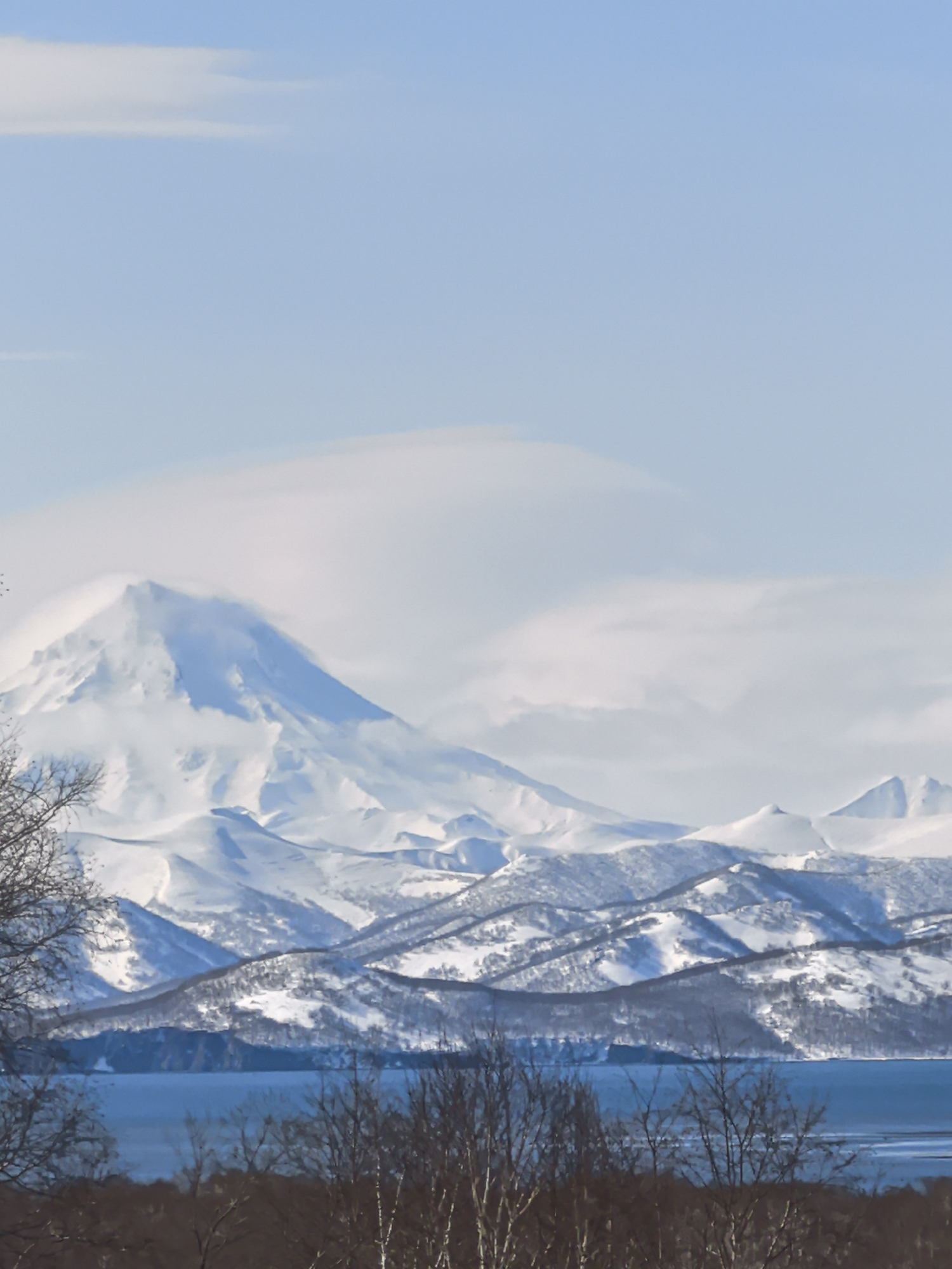 The image depicts a landscape featuring snow-covered mountains, a body of water, and trees in the foreground. A large, prominent mountain with a snow-covered peak is partially obscured by clouds. Several other mountains and hills, also covered in snow, extend across the middle ground. A body of water stretches between the mountains and the foreground, showing a darker blue hue. Bare trees and brush occupy the foreground, with their dark branches contrasting against the lighter tones of the mountains and sky. The sky is a light blue with some scattered clouds. The location is indicated as Unknown, Unknown.