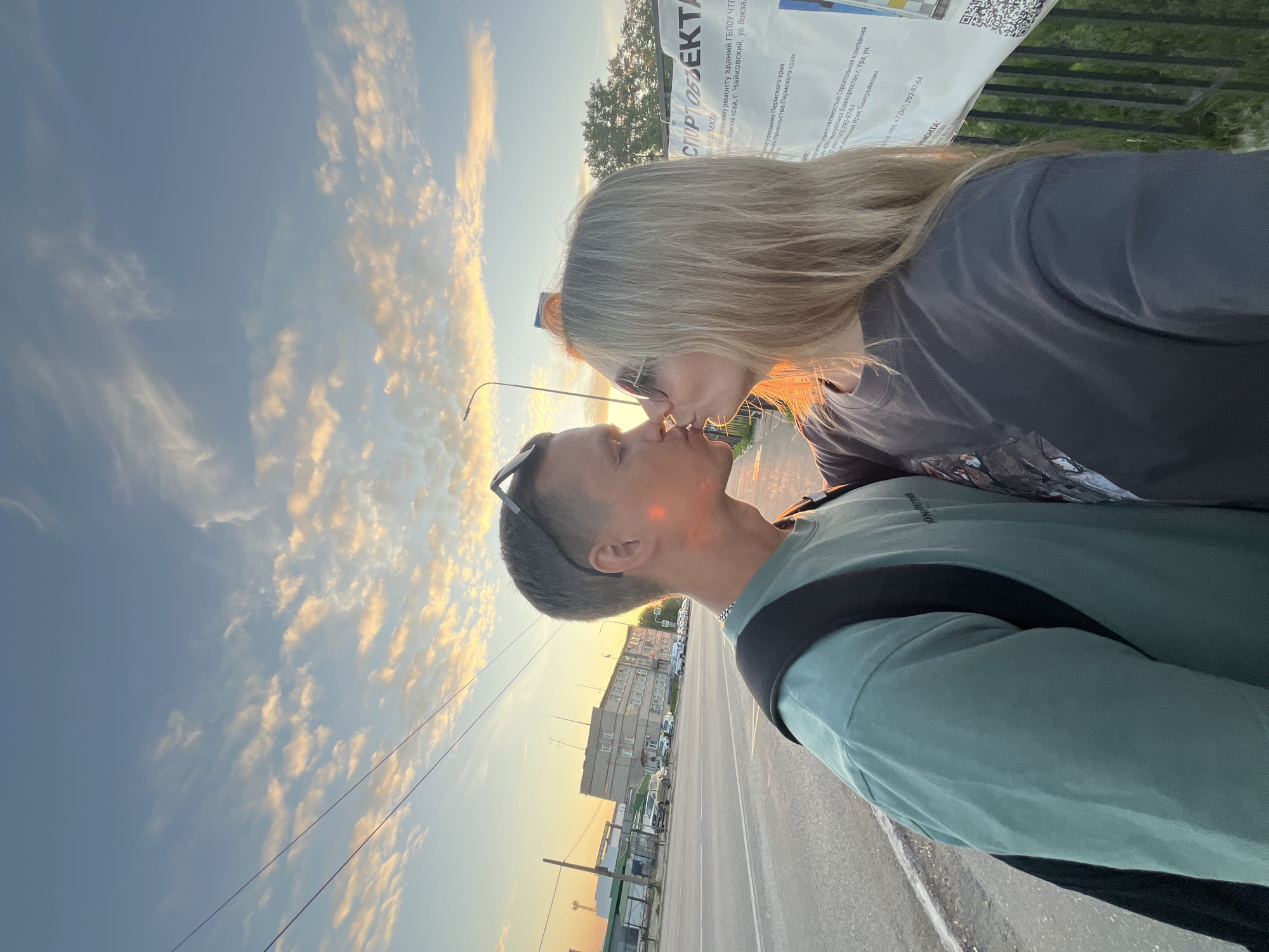 Two individuals, a male and a female, are depicted kissing outdoors at sunset in Chaikovsky, Russia. The male, positioned on the left, has short dark hair with sunglasses resting on his head. He wears a teal long-sleeved t-shirt, a chain around his neck, and a black backpack. The female, on the right, has long blonde hair and wears sunglasses over her eyes, a dark grey long-sleeved t-shirt. Both individuals are illuminated by warm, golden light on the sides of their faces closest to the camera.
They are standing near a paved surface, possibly a sidewalk, next to a road that extends into the background. Utility wires cross the upper portion of the frame. Across the road, several multi-story buildings are visible, including one light-colored building. Vehicles are parked along the roadside in front of these buildings, one appearing to be a police car. A street light pole is visible behind the couple. Behind the female, a black metal fence and a large white banner are present. The banner displays text, including "СПОРТ ОБЪЕКТА" and "г. Чайковский," confirming the location in Perm Krai, Russia. A QR code is visible on the bottom right of the banner. The sky features scattered clouds, with their undersides illuminated by the setting sun, creating warm orange and yellow hues against a light blue backdrop.