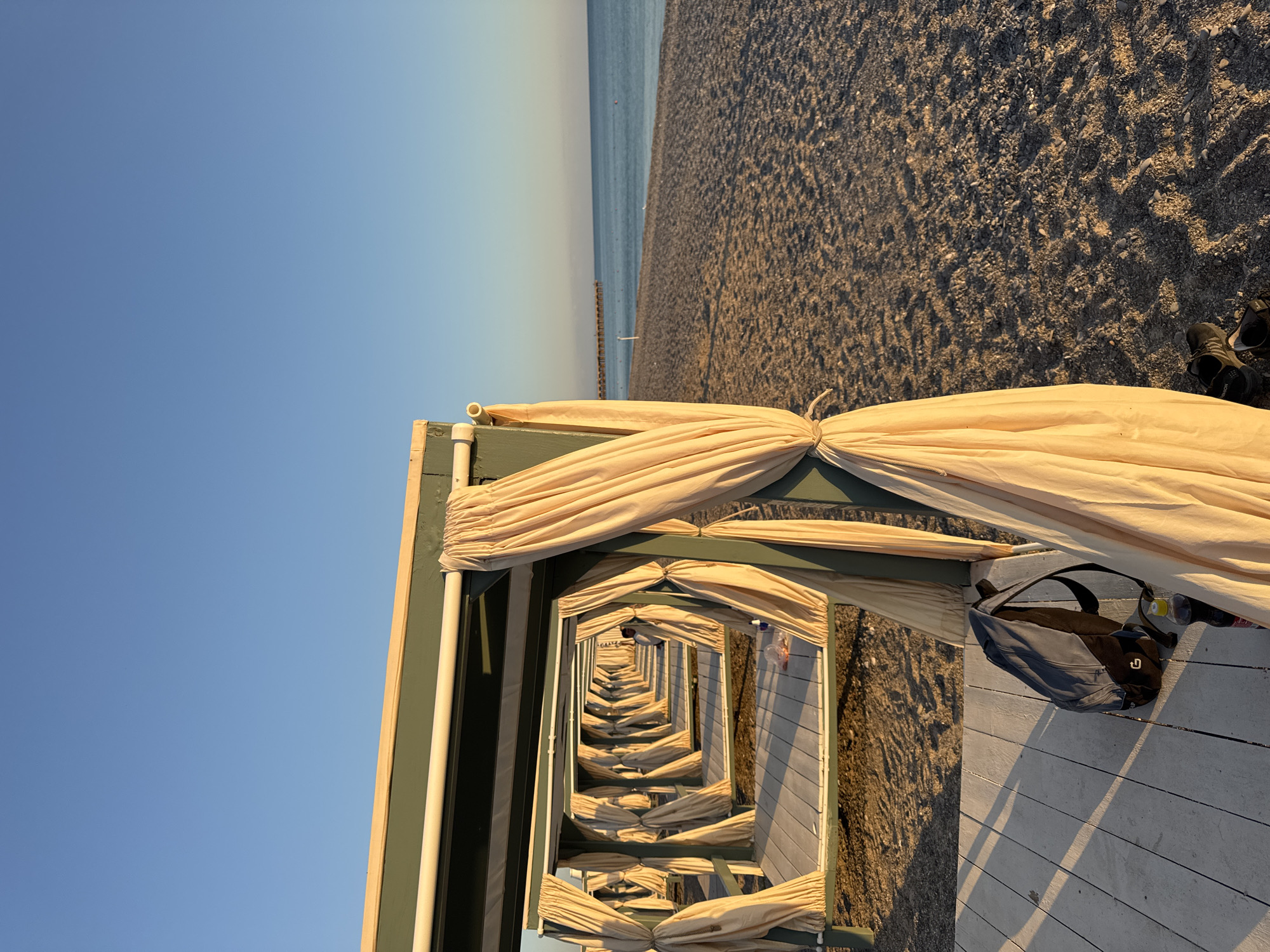 The image depicts a row of open-air cabanas on a beach, possibly located in Novofedorovka, Ukraine. The cabanas have wooden platforms and are draped with light-colored curtains. The beach is covered in pebbles and sand, and the sea is visible in the background, stretching towards the horizon under a clear blue sky. Sunlight casts long shadows across the wooden platforms, indicating it is either early morning or late afternoon. A backpack and a plastic bottle are visible on the platform of the closest cabana. A person can be seen in the distance, standing within one of the further cabanas. The scene suggests a peaceful, seaside setting.