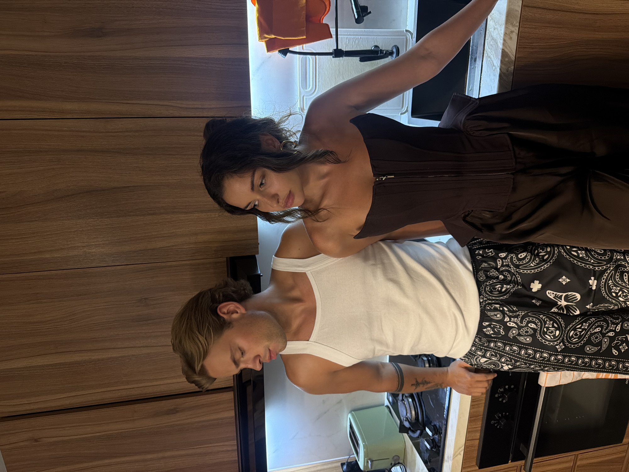 A man and woman stand in a kitchen, likely indoors. The woman, with dark hair pulled back in a bun, wears a brown corset-style top with a full skirt. She stands near a sink with a dish rack containing orange fabric. The man, with short blonde hair, wears a white tank top and black bandana-print shorts. He stands near a gas stovetop and an oven. A small, green toaster oven is visible next to the stove. The kitchen features wood cabinets and white countertops. The photo does not provide enough information to place the location in Kerobokan, Indonesia.