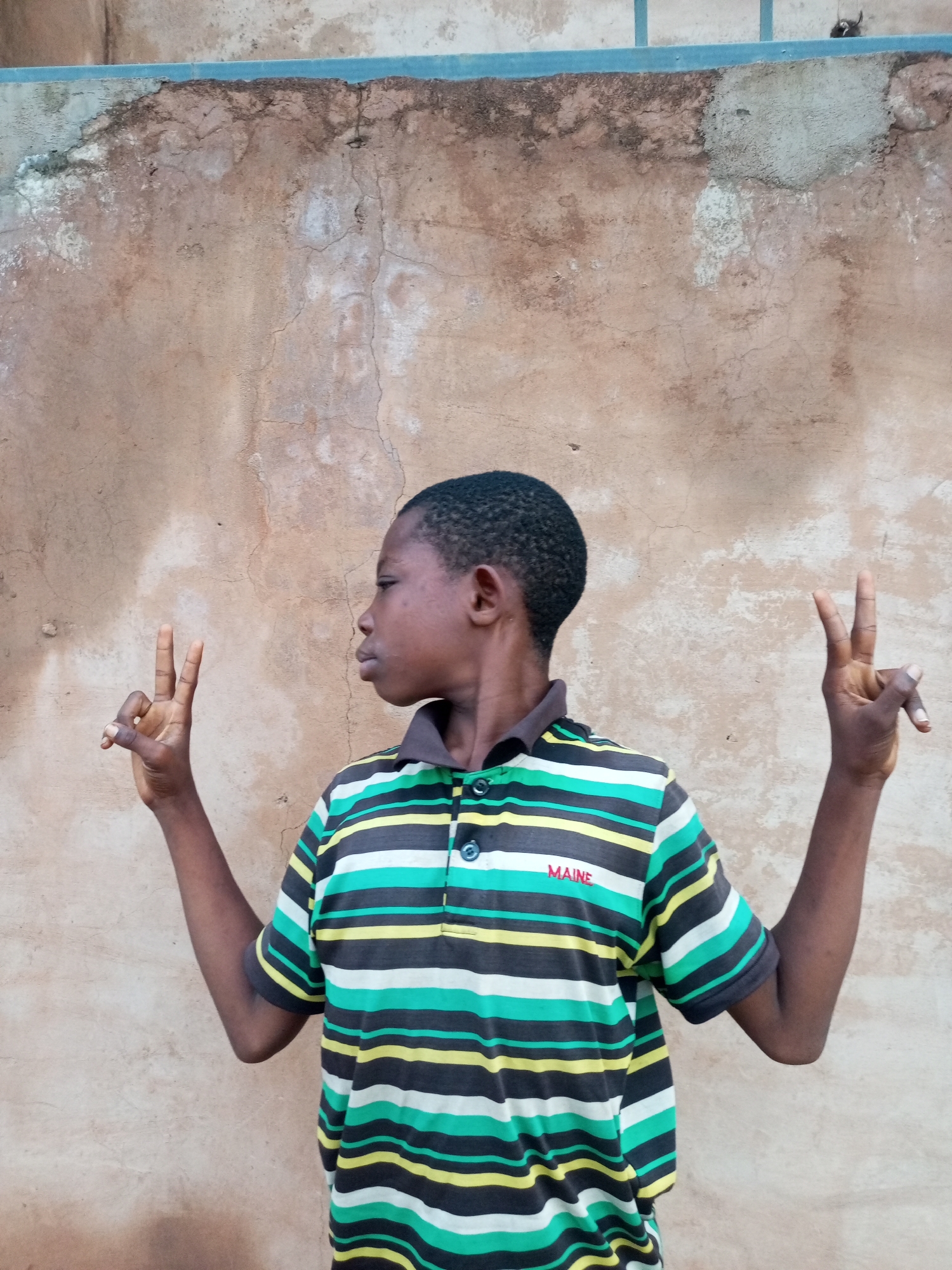 A young boy, appearing to be of African descent and around 10 years old, is standing in front of a textured, light brown wall. He is facing to his left, with his head turned slightly to his right. He has short, dark hair. He is wearing a collared shirt with horizontal stripes in black, white, green, and yellow. On the left side of his chest, the word "MAINE" is embroidered in red.
The boy is holding up both of his hands, with his index and middle fingers extended in a peace sign on each hand. His left arm is bent at the elbow and held out to his side, while his right arm is also bent at the elbow and extended outwards. The wall behind him shows signs of age and weathering, with cracks and patches of lighter color. The lighting suggests it is daytime, with the sun casting soft shadows. There are no other people or objects clearly visible in the immediate vicinity. The scene is outdoors, likely in a residential or public area.