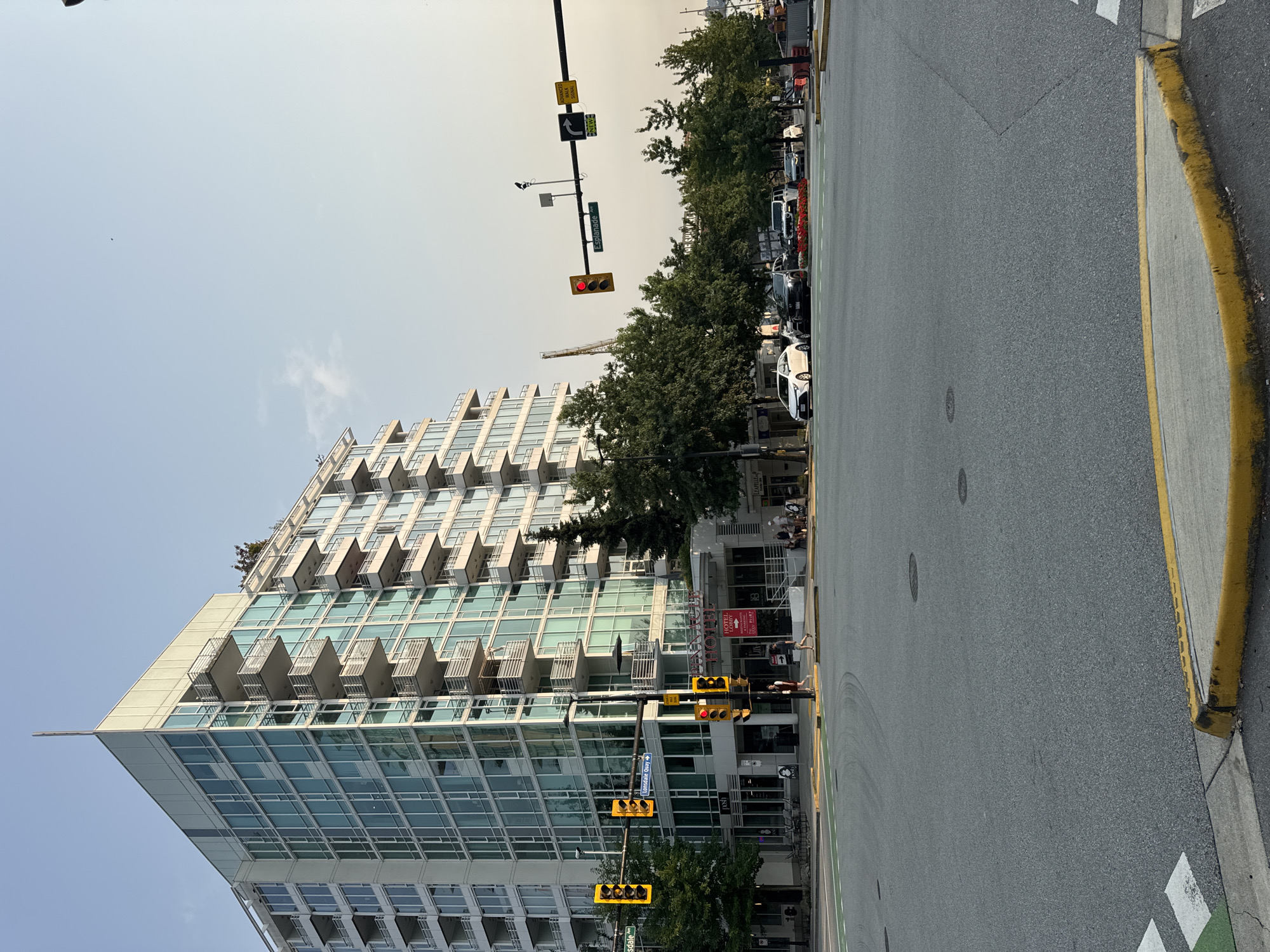 An aerial photograph captures a street scene in North Vancouver, Canada, featuring a modern, multi-story building with glass facades and balconies. In the foreground, a street intersection with visible lane markings and a yellow curb dominates the lower portion of the frame. Traffic lights are visible, with one showing a red light. Several cars are present on the street, some of which are stopped at the intersection. Pedestrians can be seen on the sidewalks and crossing the street. Trees line the sidewalks, providing greenery. A building with a "HOTEL LOBBY" sign is also visible, integrated into the ground floor of the larger structure. The sky is clear with a light haze.
