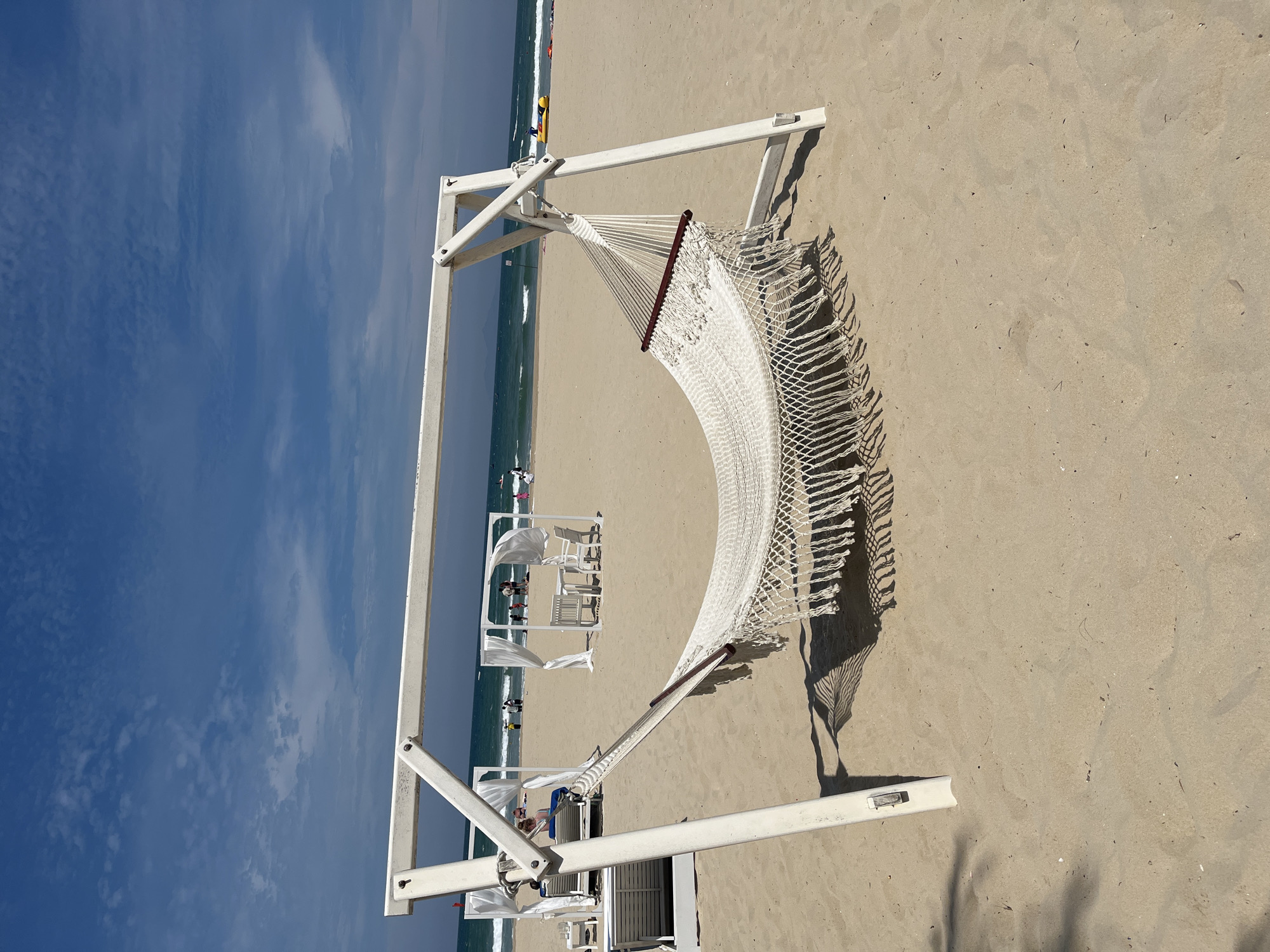 This image depicts a tranquil beach scene, likely set in Đà Nẵng, Vietnam, as indicated by the location context. The prominent subject is an empty white rope hammock, featuring a dark wooden spreader bar, suspended from a sturdy white metal stand. This setup occupies the right side of the frame, resting directly on the light beige sand. The hammock and its stand cast sharp, intricate shadows onto the sand, suggesting bright sunlight typical of midday or early afternoon.
To the left, the expansive blue ocean meets the sandy shore with gentle, breaking waves. In the mid-ground, along the coastline, several white resort-style structures are visible, including open-sided cabanas or covered lounge areas furnished with white chairs and tables. A few individuals are scattered on the beach and near the water's edge, seemingly engaged in typical leisure activities, though their actions are indistinct. A small yellow and blue object can be seen in the water close to the shore.
The sky overhead is a clear, vibrant blue, punctuated by sparse, wispy white clouds. The overall impression is one of peace, relaxation, and an idyllic vacation setting under excellent weather conditions. There is no visible text present in the image.