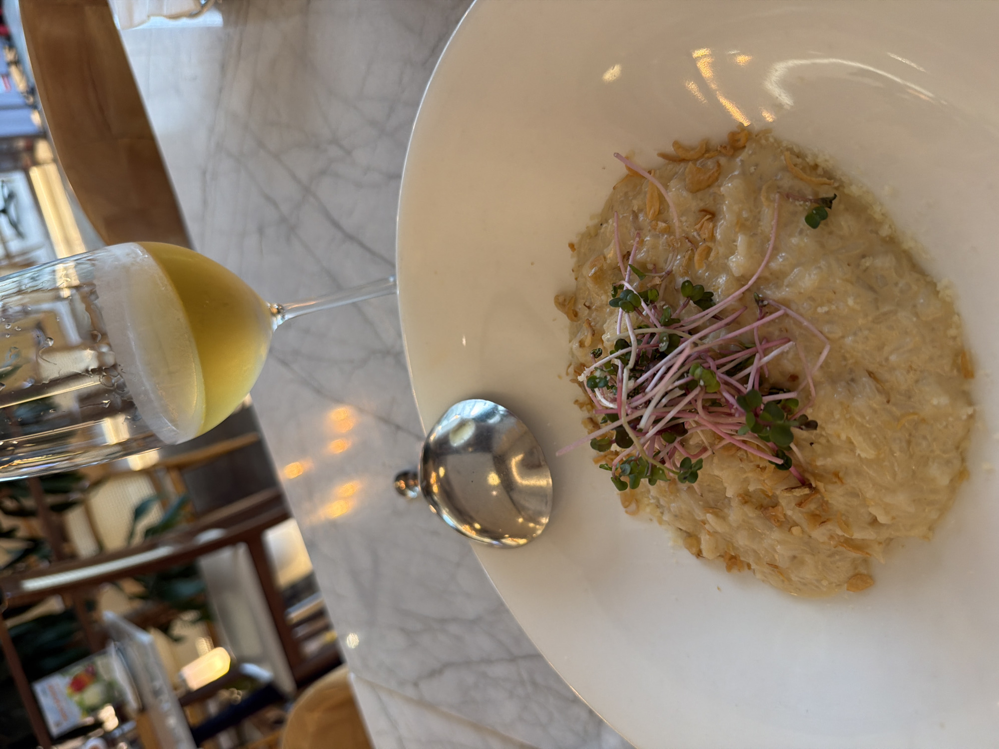 A close-up, overhead shot showcases a bowl of risotto and a glass of yellow liquid on a marble table. The risotto is a creamy, pale yellow dish topped with thin, crispy fried shallots and a garnish of pink-stemmed microgreens. A silver spoon rests on the side of the white bowl. To the left, a wine glass contains a pale yellow beverage, possibly a white wine or juice, with condensation on the outside. The background is blurred, but suggests an indoor setting with warm lighting and wooden furniture, possibly a restaurant in Hanoi, Vietnam. The lighting indicates it could be daytime.