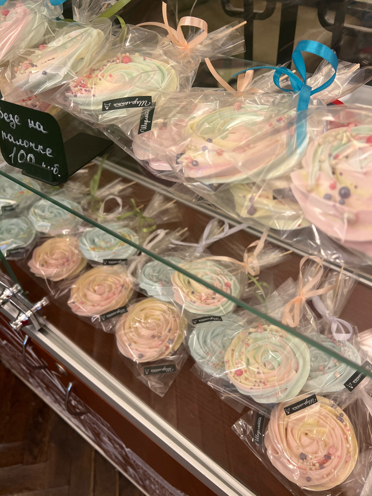 The image shows a display case filled with individually wrapped meringue cookies, also known as "beze" in Russian. The cookies are pastel-colored and decorated with sprinkles. Some are on sticks, resembling lollipops, while others are laid flat. A handwritten sign in Russian is visible, with text that appears to be "Безе на палочке 100 руб." which translates to "Meringue on a stick 100 rubles." This suggests the location is a shop or bakery selling sweets. The lighting is bright, and the overall scene is clean and well-organized. The colors of the meringues are soft and appealing, and the clear plastic packaging highlights the delicate nature of the confectionery. The image is taken from a slightly elevated angle, looking down into the display case. The background is blurred but appears to be the interior of a retail establishment. There are no people or animals visible in the image. The context of Kaliningrad, Russia, is not explicitly evident from the visual information alone but is provided as external information.