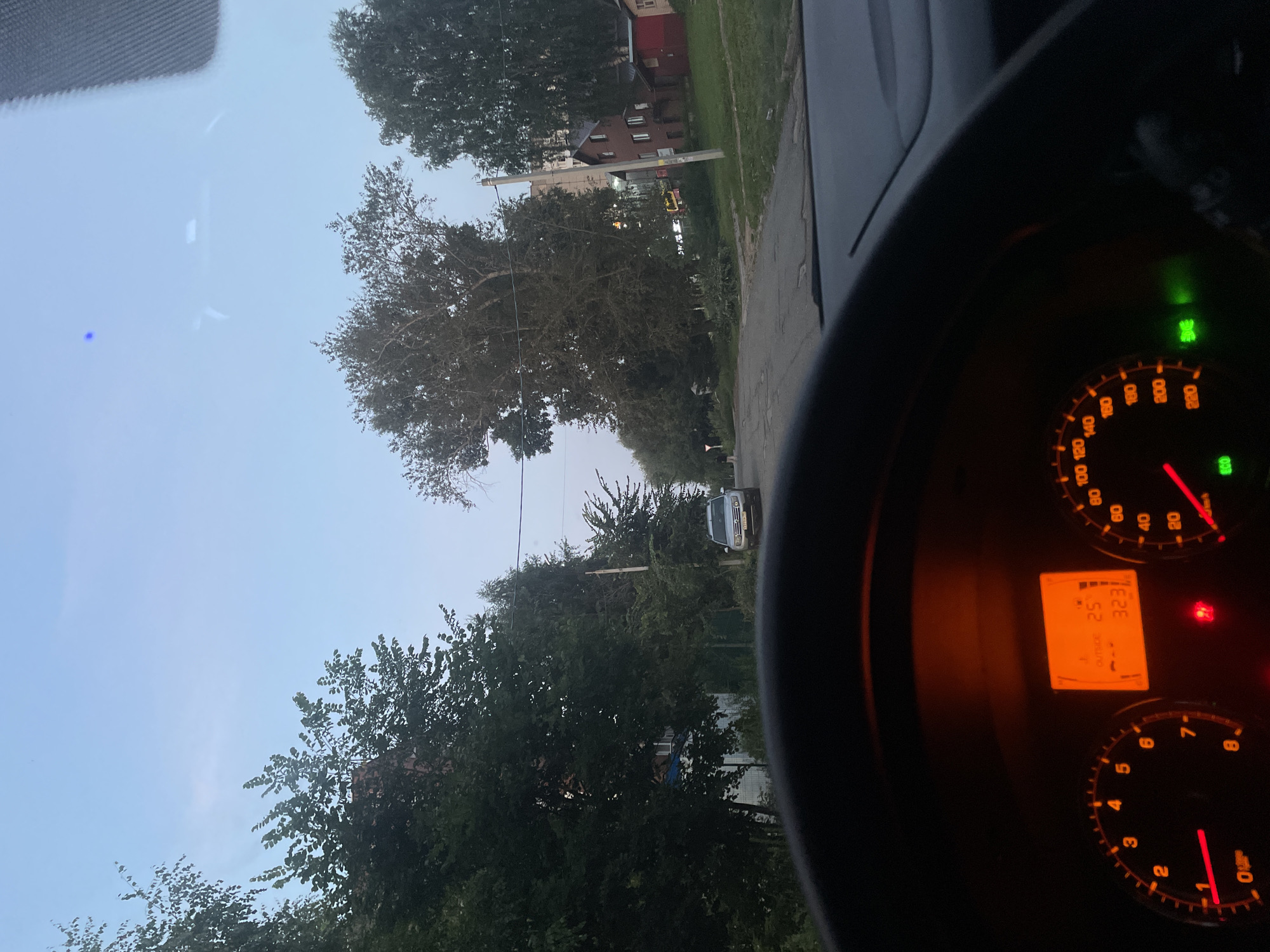 The image shows a view from the driver's seat of a car, looking forward through the windshield. The steering wheel is visible in the foreground, with illuminated dashboard instruments. The instrument panel displays an outside temperature of 25 degrees Celsius and a mileage of 3231. The car is on a paved road in a rural or suburban area. Tall trees line both sides of the road, and there are houses and buildings visible in the background. The sky is clear and pale blue, suggesting it is either early morning or late evening. A silver SUV is parked on the right side of the road. In the distance, a white traffic sign can be seen. The environment appears to be Votkinsk, Russia, given the overall landscape and typical architecture.