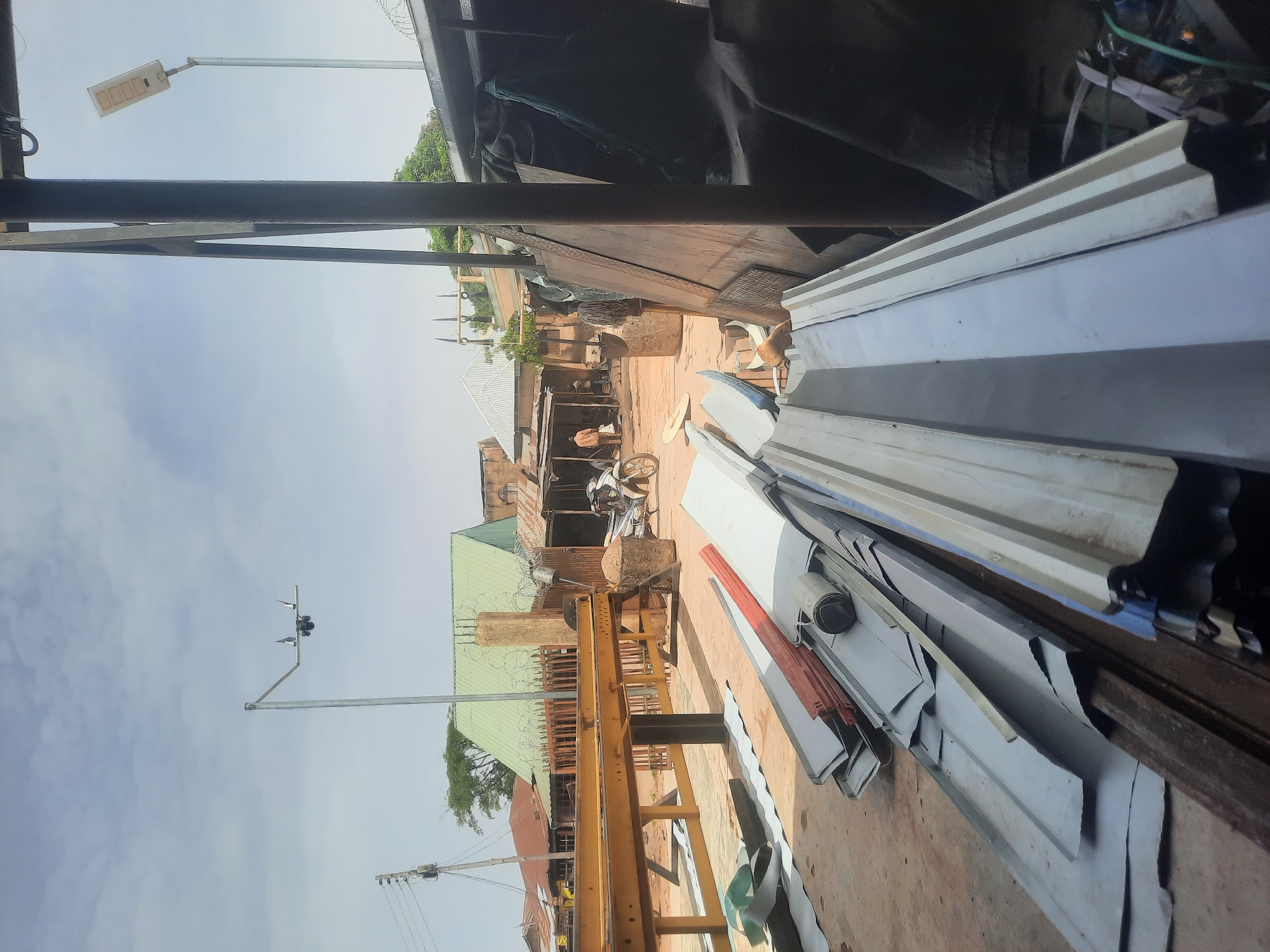 The image depicts a street scene in Gura Top, Nigeria, with various structures and objects in view. A man on a bicycle is present. There are houses, some with green roofs, and light poles in the distance. In the foreground are stacked corrugated metal sheets and a yellow metal frame structure. The environment appears to be a combination of residential and industrial elements.