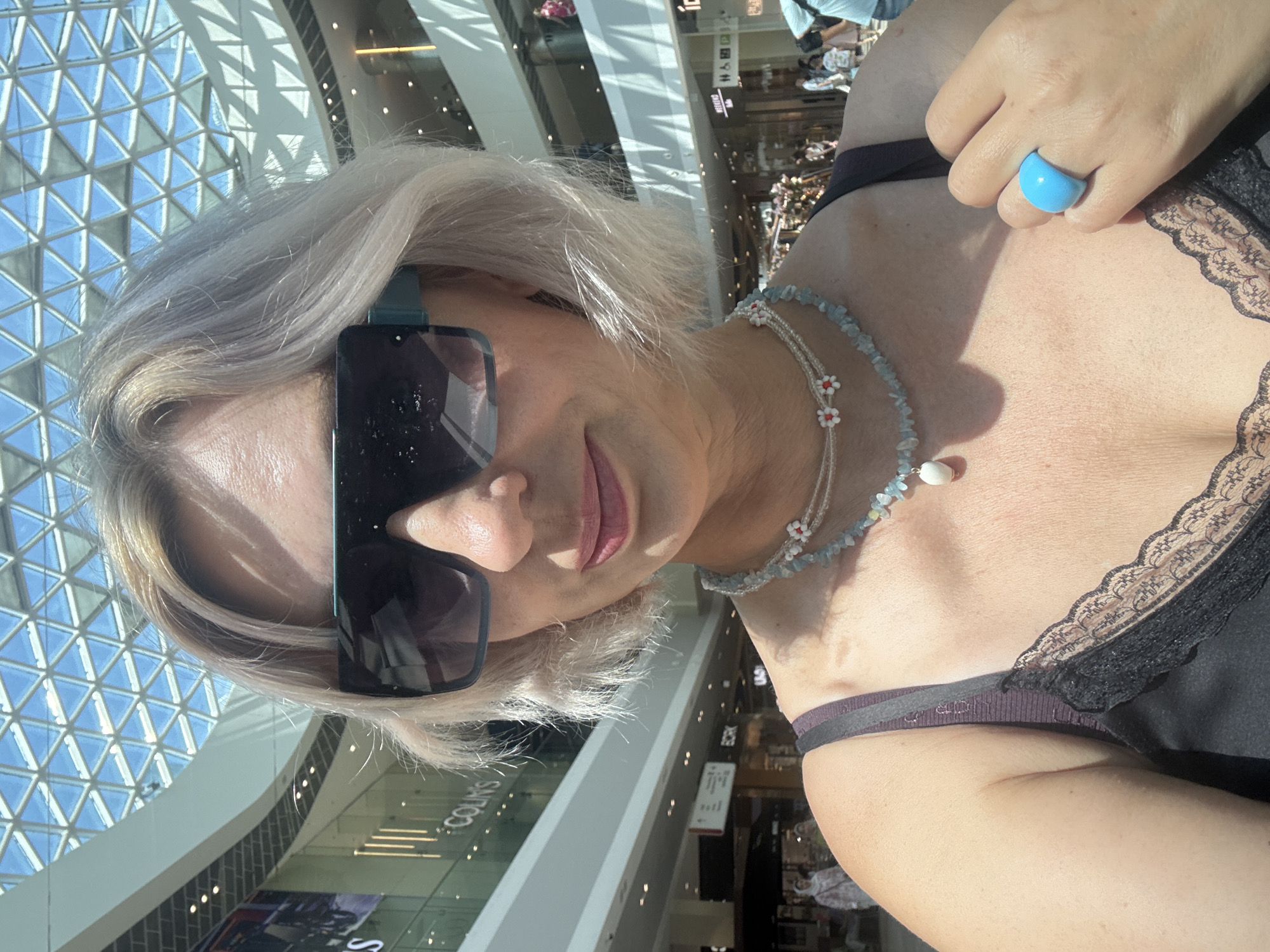 A woman with short, light-colored hair and large sunglasses takes a selfie in a brightly lit indoor shopping mall. She is wearing a black camisole with lace trim and several necklaces. One necklace features light blue beads and a white pendant, while another is a delicate white string adorned with small red and white floral beads. She also wears a bright blue ring on her right index finger. The background shows the multi-level architecture of the mall, with a large glass ceiling, escalators, and shop facades. The environment appears to be modern and spacious, with natural light filtering through the roof. The woman's expression is neutral as she captures the photo. The image suggests a casual outing in a commercial setting. The specific location is identified as a shopping mall in the city of Sochi, Russia.