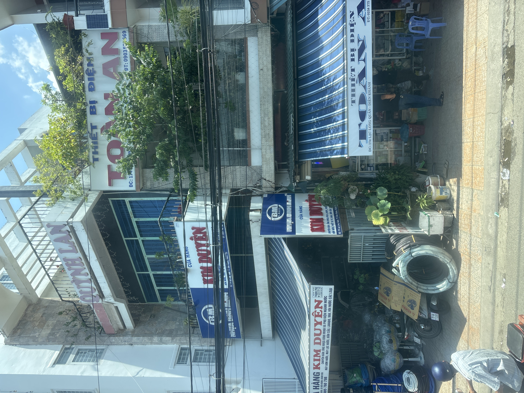 The image shows a street scene in a city with multi-story buildings. The buildings are commercial establishments, as indicated by the signs and awnings. One prominent sign reads "QUỲNH LAN" in large red letters on the upper floor of a building. Below this, another building features a sign for "THIẾT BỊ ĐIỆN TOÀN AN". Various signs for "CỬA HÀNG KIM DUYÊN" are also visible, suggesting a business selling goods. In front of the shops, there are awnings providing shade, merchandise such as large coils of tubing, fans, and a parked motorcycle. People are present, including individuals standing inside a shop and a person on a motorcycle in the foreground. The environment appears to be a typical urban street with overhead electrical wires and a sidewalk.