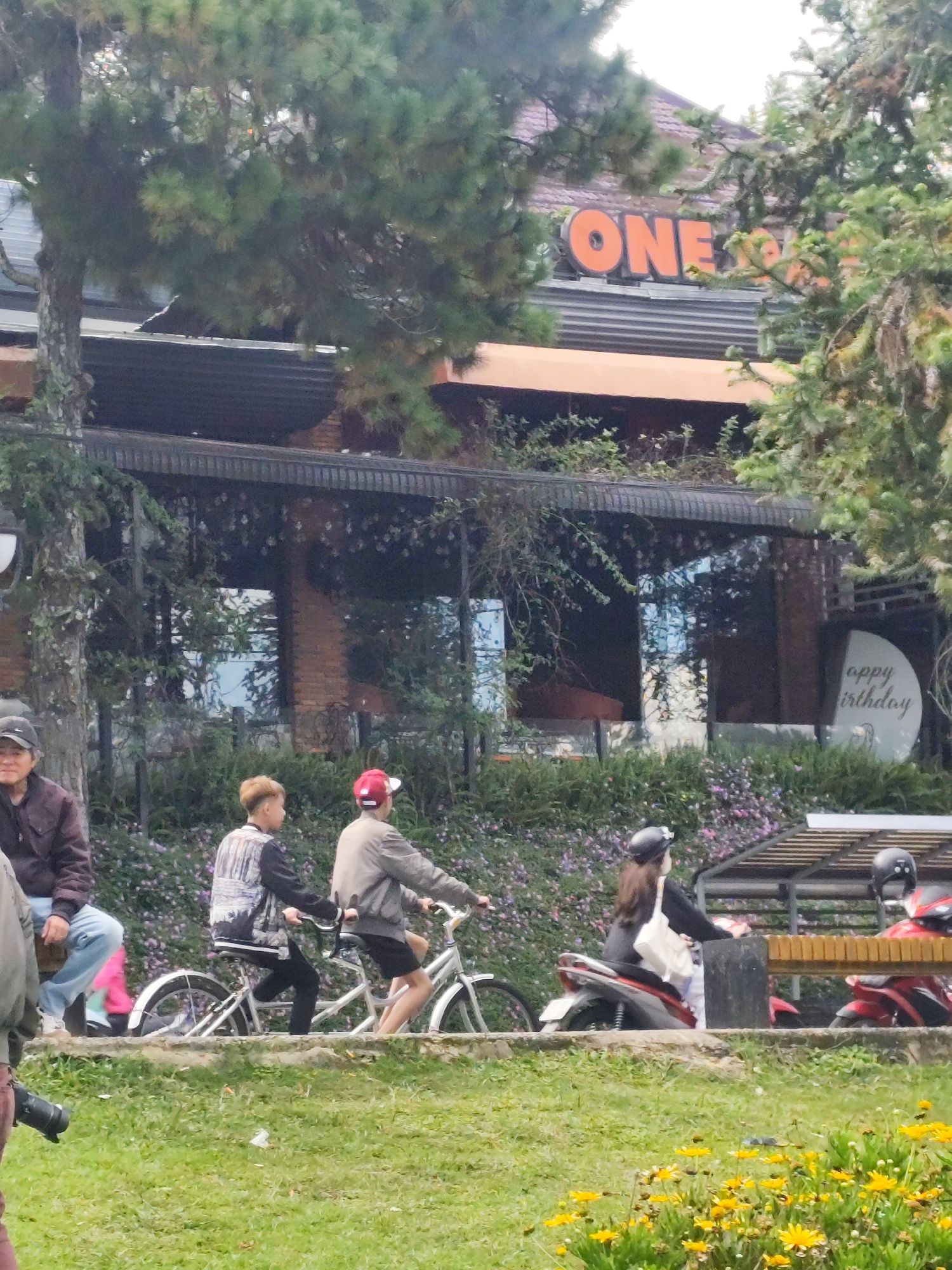 The image captures a street scene in Da Lat, Vietnam. In the foreground, a patch of grass is dotted with yellow flowers. Behind the grass, a stone wall with a grey railing runs horizontally. Two young individuals are riding side-by-side on tandem bicycles. The person on the left, a male with short blonde hair, wears a black and white patterned jacket. The person on the right, also appearing to be male, wears a light-colored jacket and a red baseball cap.
Further back, a person in a dark jacket and light blue jeans sits on the stone wall, facing left, partially obscured by a tree. To the right of the cyclists, a young woman wearing a black top, helmet, and a white tote bag is riding a scooter, also facing right. Behind her, another scooter with a red body is parked.
In the background, a building with a brick facade is visible. Orange letters spelling "ONE" are prominently displayed on the upper part of the building. A circular sign with white text, partially readable as "appy irthday," is affixed to the building's glass facade. Trees with dense green foliage partially obscure the building and the sign. The overall atmosphere appears to be a casual, outdoor setting in a city, possibly a cafe or a commercial establishment given the signage.