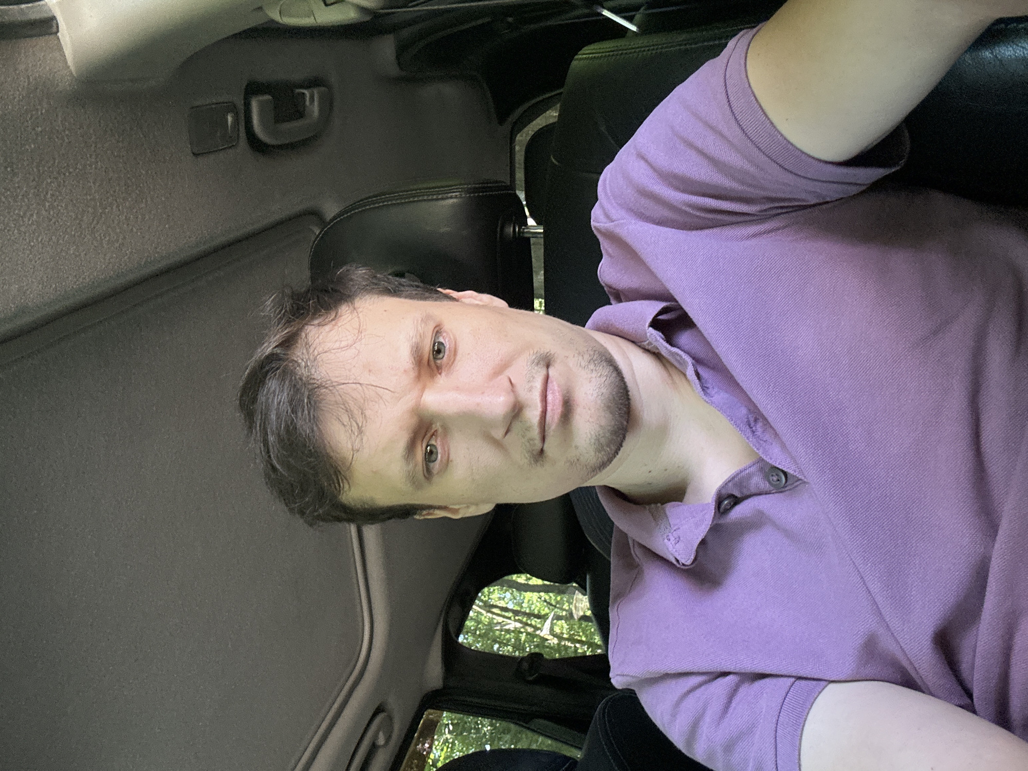 The image shows a medium shot of a man in a purple polo shirt sitting in the driver's seat of a car. He is looking directly at the camera with a neutral expression. The car's interior is visible, including the headrest of the passenger seat and a sunroof. Through the windows, lush green trees and foliage are visible, suggesting an outdoor, possibly wooded, location. The man's skin tone is light, and he has short, dark brown hair. The lighting is natural and appears to be daylight. There is no indication that this scene is located in Shatava, Ukraine.