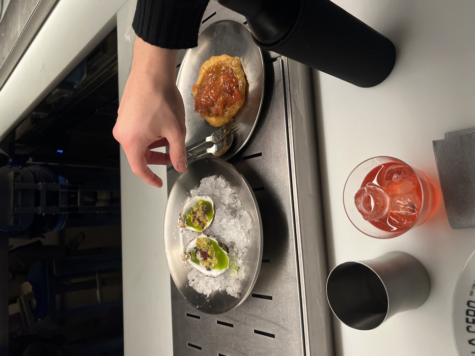 A hand hovers over a plate with two oysters on crushed ice, ready to pick one up. Above that plate is another with a small tart. A water bottle sits next to the tart plate. A glass of a red beverage with ice, a metallic shaker, and a plate with a logo are on the table as well. The scene is set on a metallic surface next to a white table.