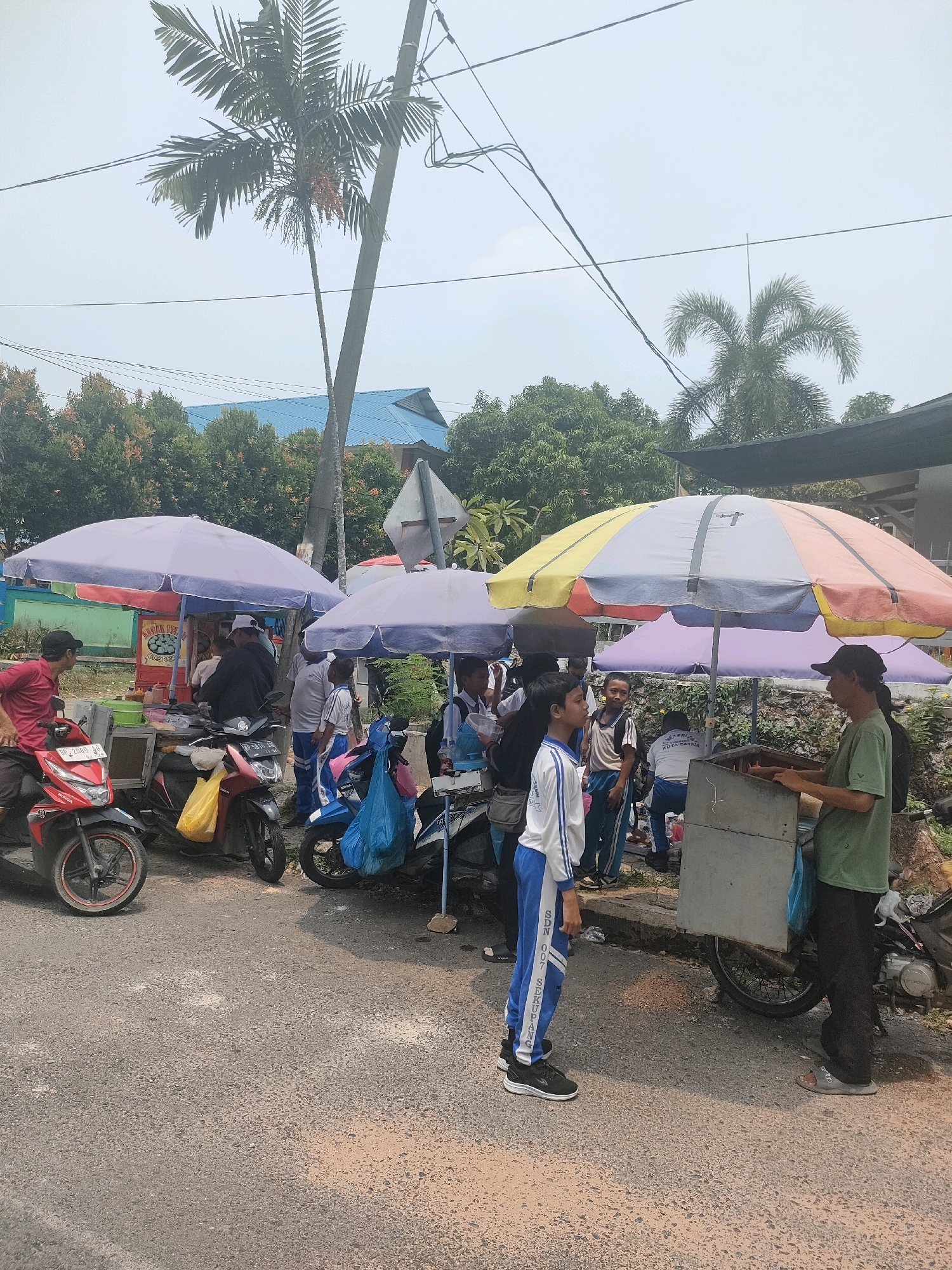 A daytime scene depicts an outdoor street food vendor area in Batam, Indonesia. Several food carts, covered by colorful umbrellas, are lined up along a paved road. People, including children in school uniforms and adults, are gathered around the carts, some appearing to be customers or vendors. Motorbikes are parked nearby. Palm trees and other foliage are visible in the background, along with a building with a blue roof. The sky is overcast.
