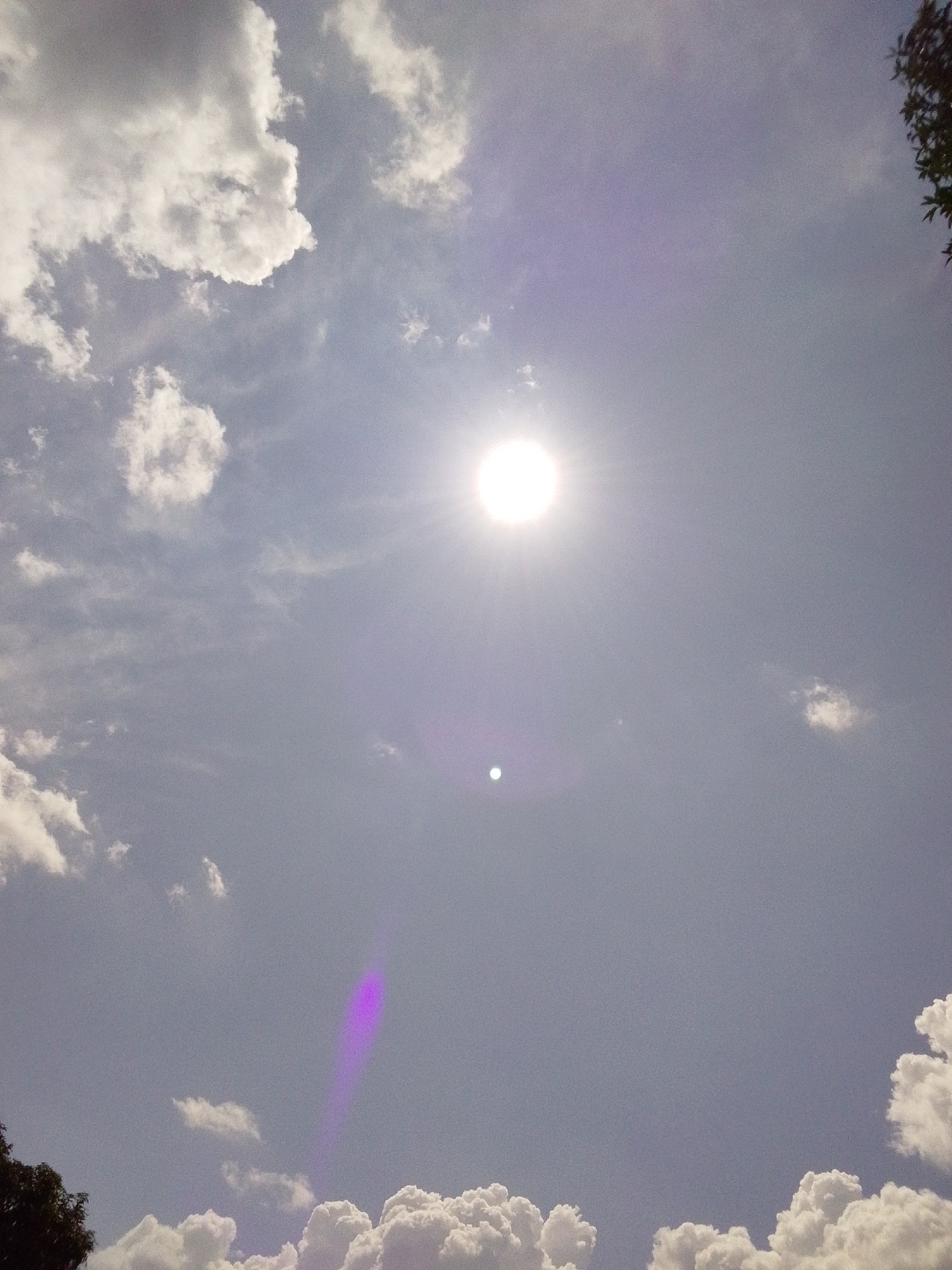 The image shows a bright sun in a hazy sky with scattered clouds. The sun is positioned in the upper center of the frame, emitting a strong glare. There are various cloud formations visible, ranging from thin wisps to larger, more dense cumulus clouds. The overall impression is of a sunny day with some atmospheric haze. The provided crops do not offer any information about a city or location.