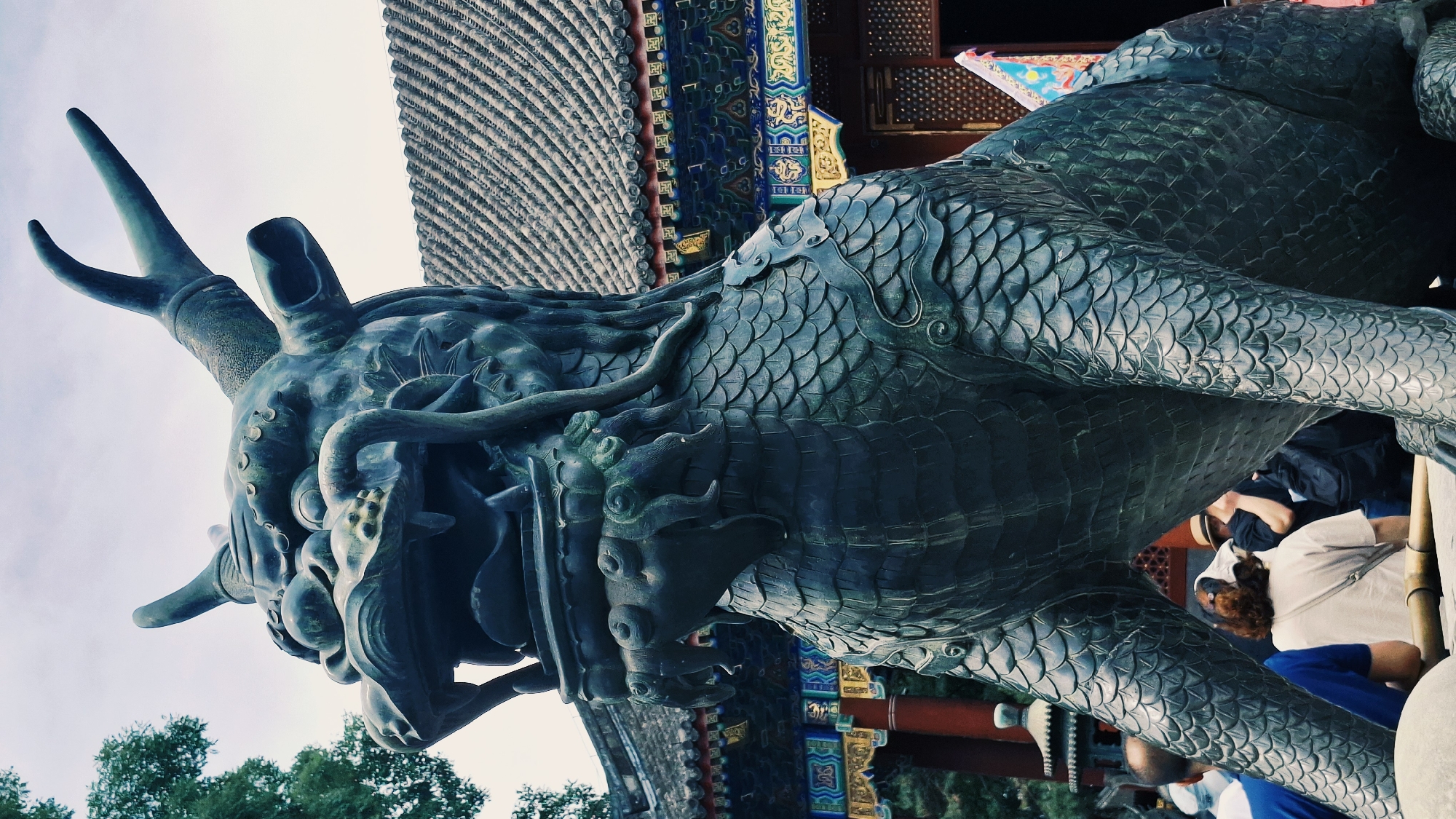 A bronze dragon sculpture, likely a public artwork or historical artifact, is depicted from a low angle against a background featuring traditional Chinese architecture and people. The sculpture, detailed with scales and ornate features, faces the viewer with the sky in the background. The architectural elements include a tiled roof and colorful geometric patterns on the building's facade, characteristic of traditional Chinese design. A small group of people is partially visible behind the sculpture, suggesting the artwork is located in a public space, possibly a park or garden. Based on the architectural style and the presence of such sculptures, the location is highly likely to be in the city of Haidian District, China.
