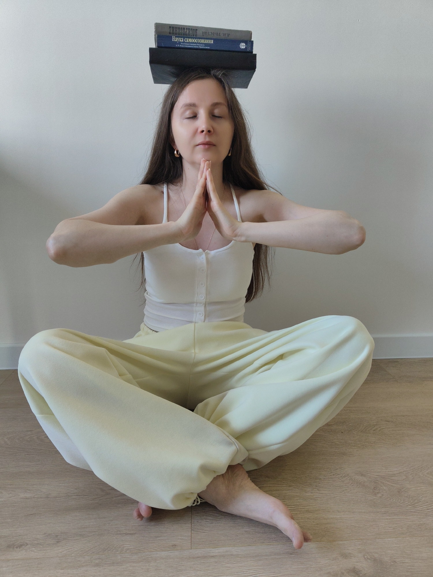 A woman sits cross-legged on a light wood floor, hands pressed together in a prayer-like gesture. She wears a white tank top and pale yellow pants. On top of her head are stacked three books. The location appears to be indoors against a light-colored wall. The city and specific location are unknown.