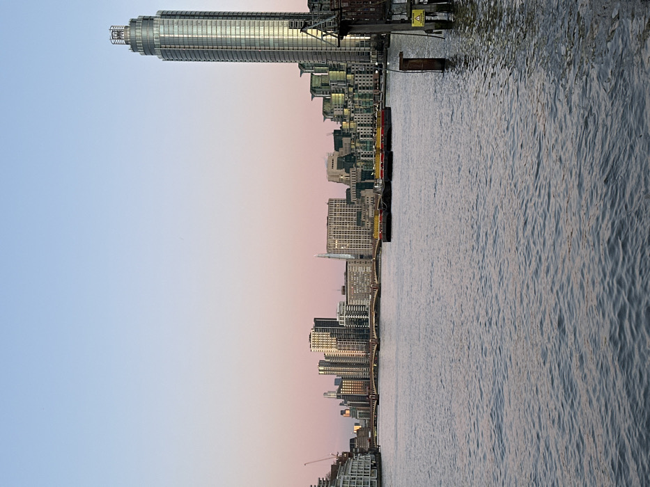 An urban skyline stretches along a body of water under a dusky sky. Tall buildings, including a prominent cylindrical skyscraper, dominate the scene. Reflections ripple on the water's surface. The sky transitions from pale blue to soft pink, suggesting twilight or sunrise. The overall scene depicts a contemporary cityscape viewed from a waterside perspective.