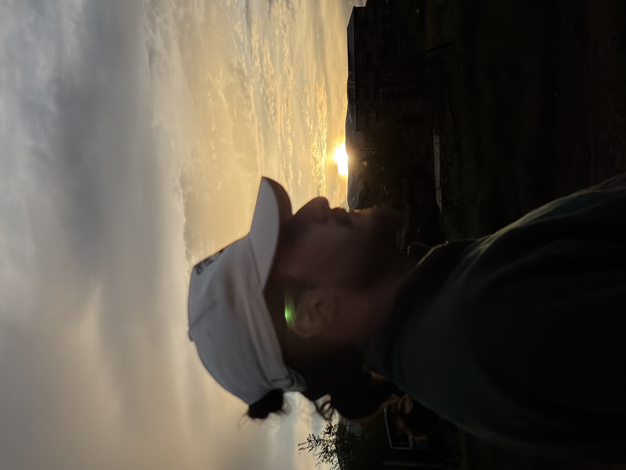 A medium shot shows a man in a green shirt and a white baseball cap looking up toward the sun. The sun is setting behind a building in the background. The photo was taken at a low angle, as if from ground level. The man is looking up, toward the sky. The photo was likely taken outdoors, during sunset. The location cannot be determined.