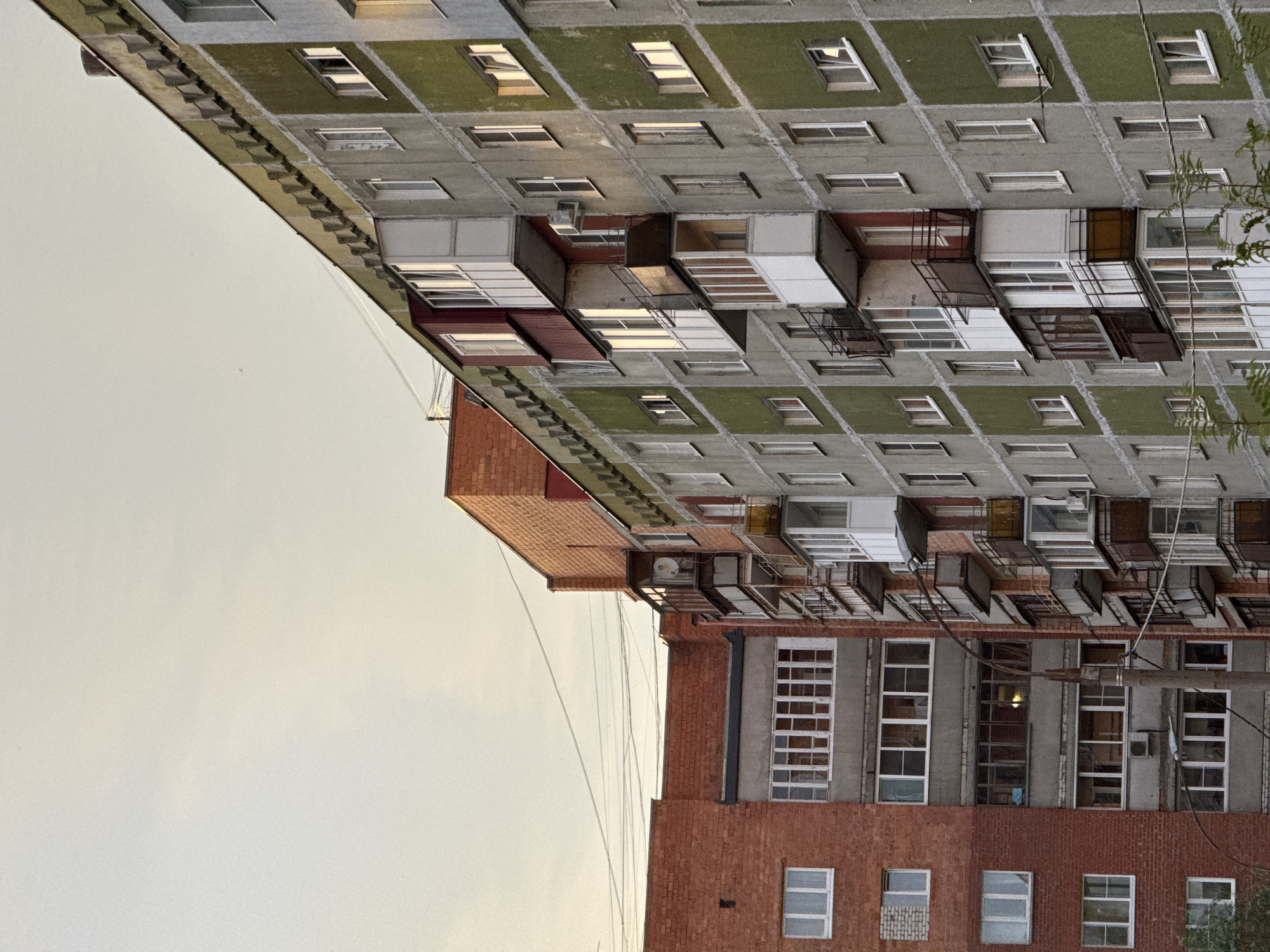 The image depicts a view of residential apartment buildings in Nizhny Novgorod, Russia, likely during the daytime with soft lighting. The architecture features a mix of brick and concrete structures with balconies and numerous windows. One building is predominantly red brick, while another is a multi-story concrete building with green panels and balconies. Electrical wires are visible crisscrossing the sky between the buildings. The scene captures a common urban landscape with a focus on the building exteriors and their structural elements. There are no people visible in the image.