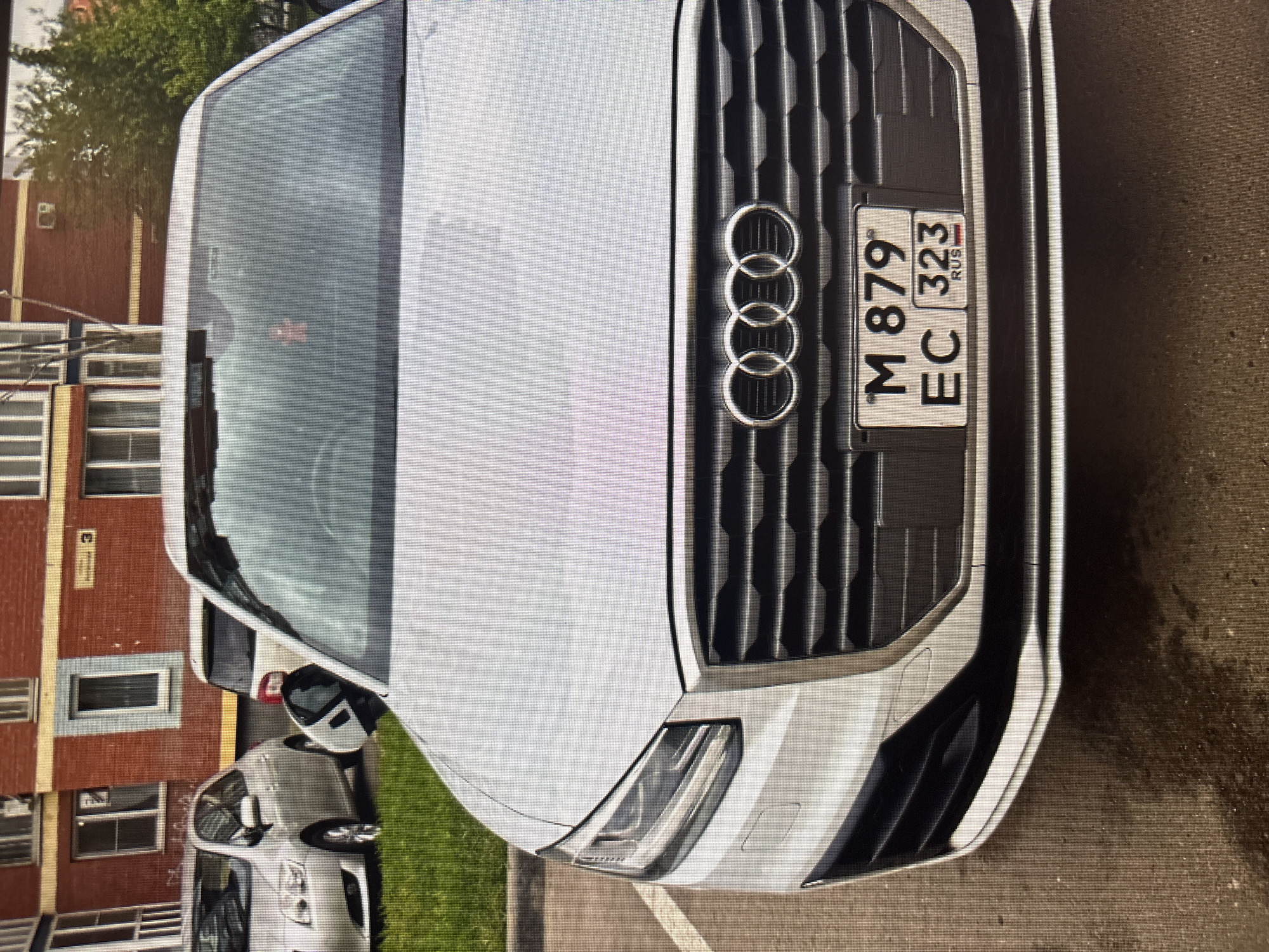 An Audi Q2 is parked in front of a brick apartment building. The car is white and has a Russian license plate that reads "M 879 EC 323 RUS". A small red figure is hanging from the rearview mirror inside the car. The building appears to be in Moscow, Russia. The foreground is paved with asphalt and there is grass to the left of the car.