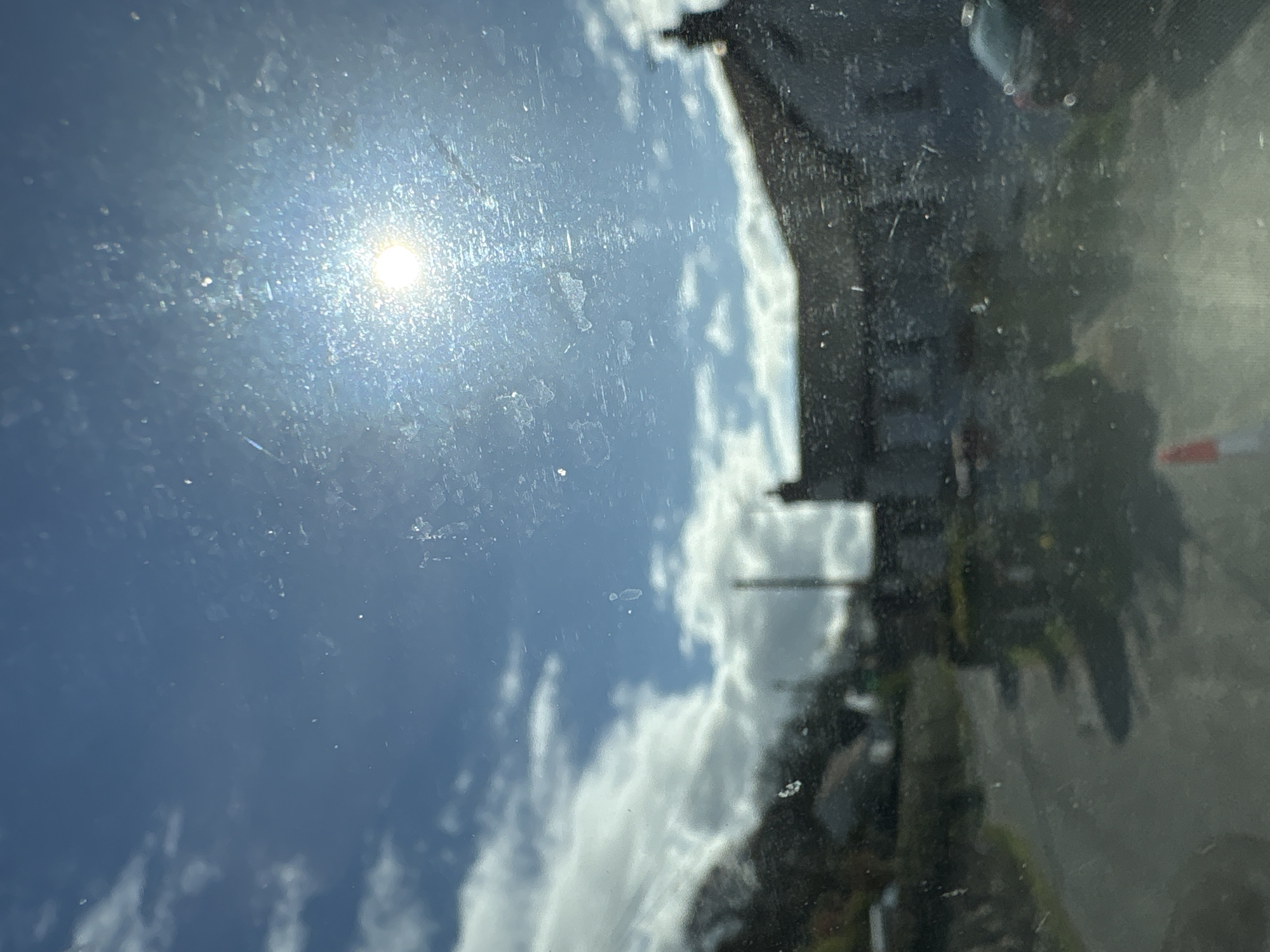 The image appears to be taken from inside a vehicle, looking outwards through a dirty or dusty window. The sun is visible in the upper left portion of the frame, obscured by the window imperfections. Reflected in the window is a scene from Snailbeach, United Kingdom, showing a building and cloudy sky. The building is dark and partially obscured. The sky is light blue with fluffy white clouds.