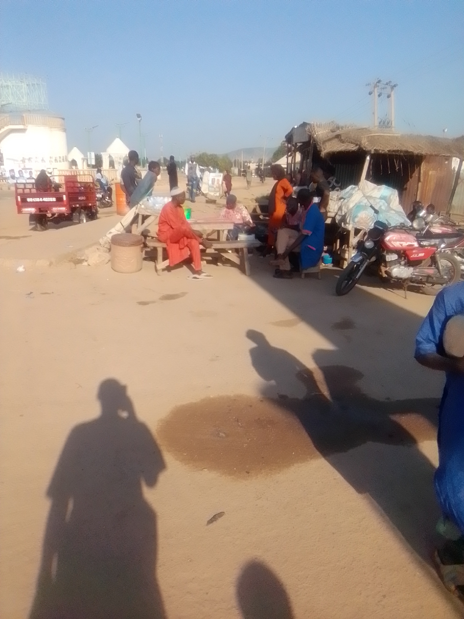 The image depicts a street scene in Kwadon, Nigeria. Several people are present, some gathered around a table, while others are standing or walking. A man in a red garment is seated on a bench. To the left, a red truck with the registration number "0838440555" and "AGBO" is parked. A motorcycle is visible in the mid-right of the image. The environment appears to be a market or a public gathering area with various structures, including stalls with thatched roofs and what looks like a building in the background. The ground is unpaved, and there are shadows cast on it, suggesting it is daytime.