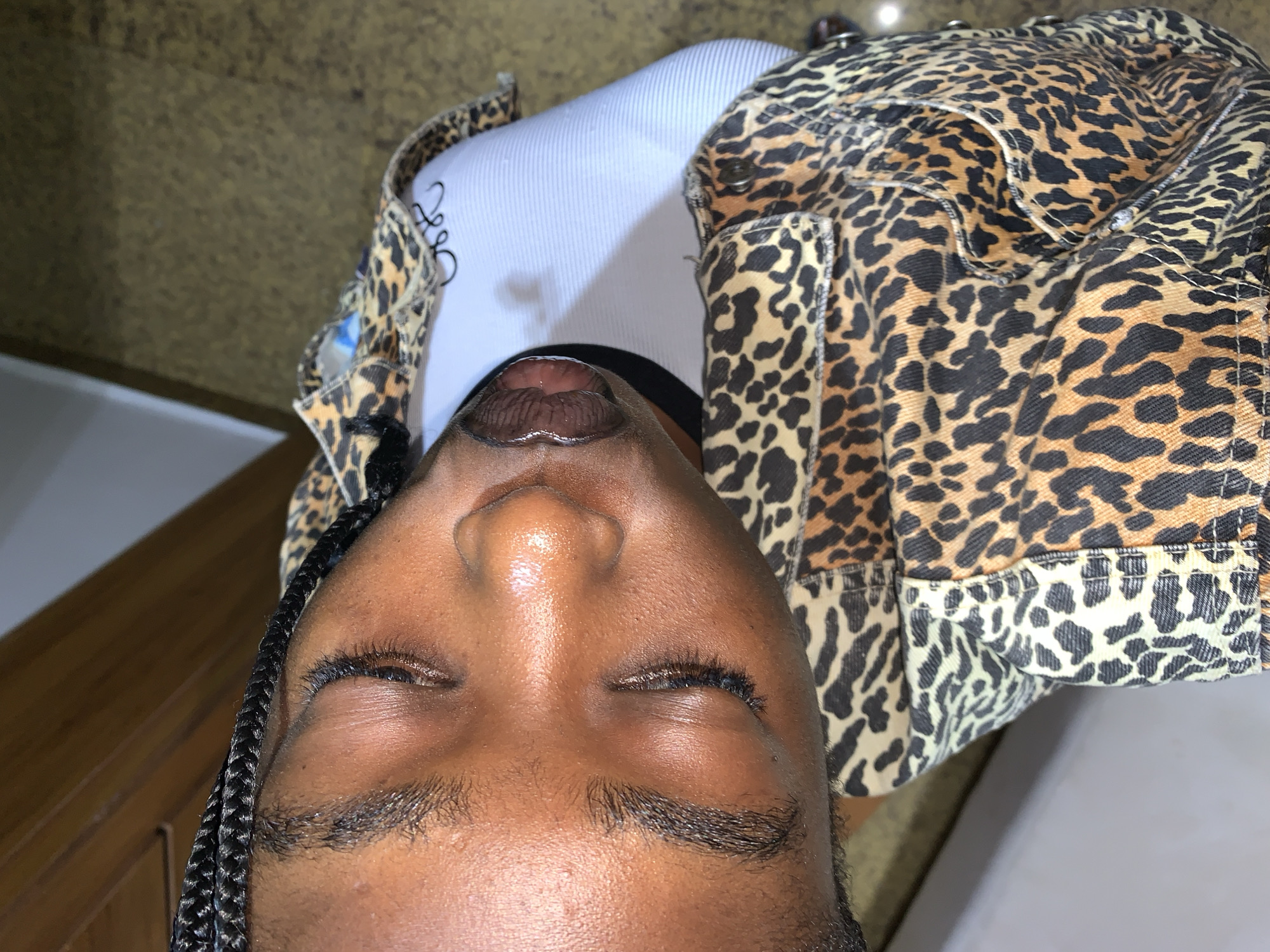The image shows a close-up, angled view of a person's face with their eyes closed. The person has dark skin and is wearing braids. They are also wearing a leopard-print jacket. The background is blurry and appears to be a textured surface. There is no indication of the specific location within Gura Top, Nigeria.