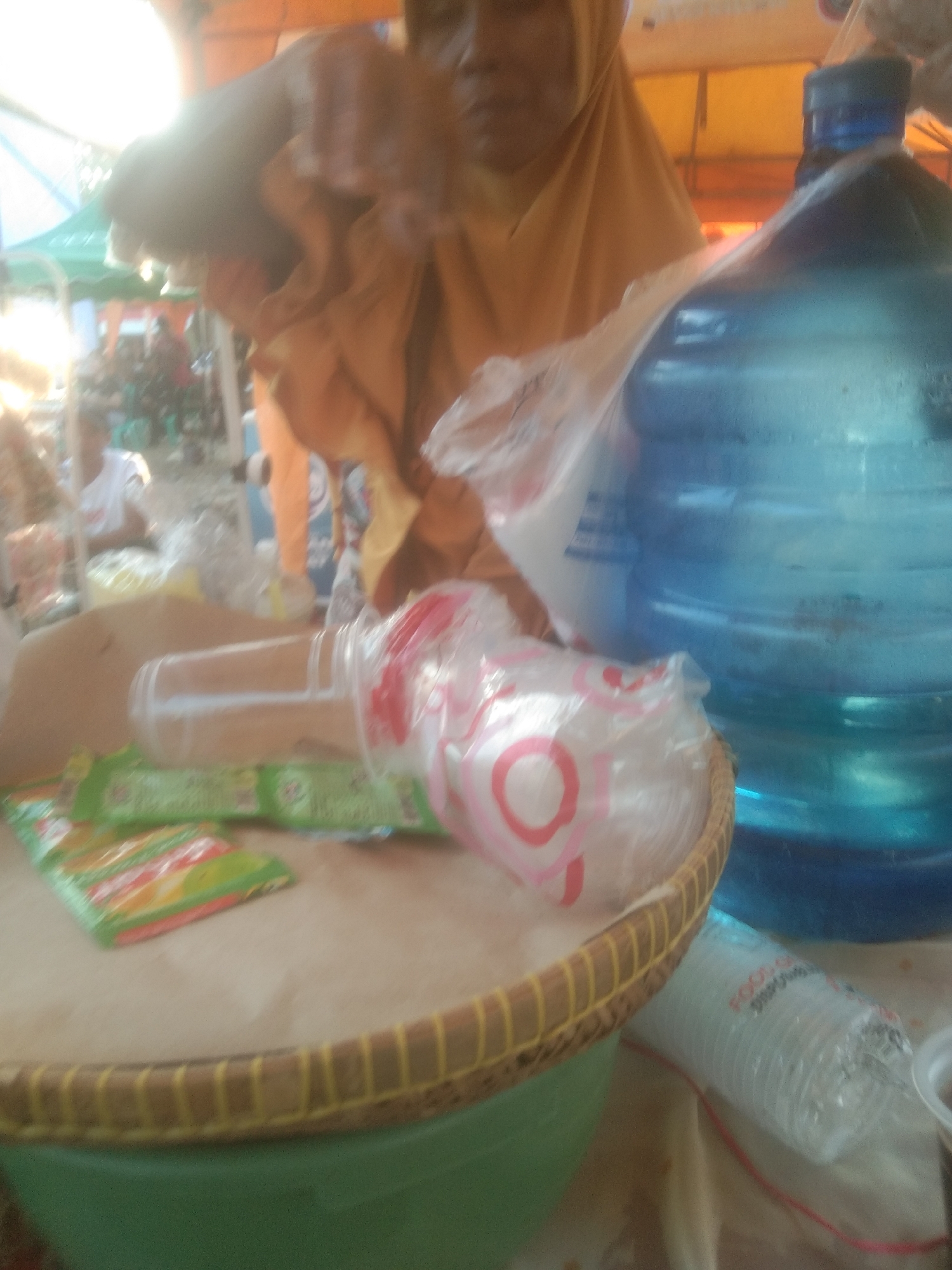 This is a photograph taken in Kecamatan Haurgeulis, Indonesia, featuring a close-up view of various items on a table outdoors, likely at a market or roadside stall. In the foreground, a woven basket holds several green-packaged food items and a clear plastic cup with pink circles. To the right of the basket is a large blue water jug with a blue cap. Behind the water jug, a person wearing a yellow headscarf is partially visible. In the background, other individuals and a green canopy are out of focus. The lighting suggests it is daytime.