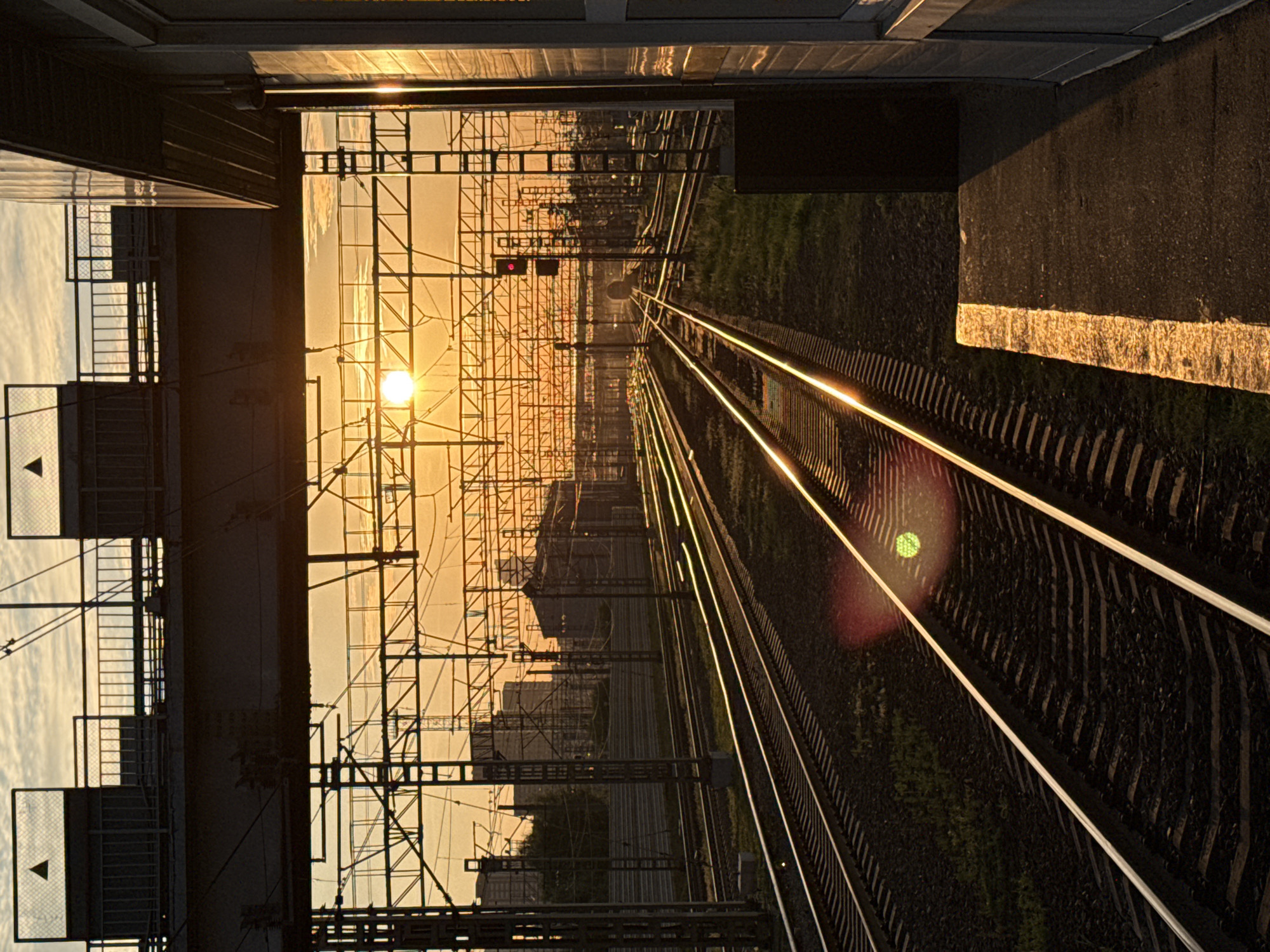 The image depicts a railway junction at sunset in Moscow, Russia. The sun is low on the horizon, casting a warm, orange glow across the sky and illuminating the complex network of overhead wires and support structures. Multiple railway tracks converge and diverge, disappearing into the distance. A pedestrian overpass spans the tracks in the foreground, with fencing and enclosed structures on top. To the right, a modern building with large windows stands adjacent to the platform. A red railway signal is visible further down the tracks. The scene is devoid of people.