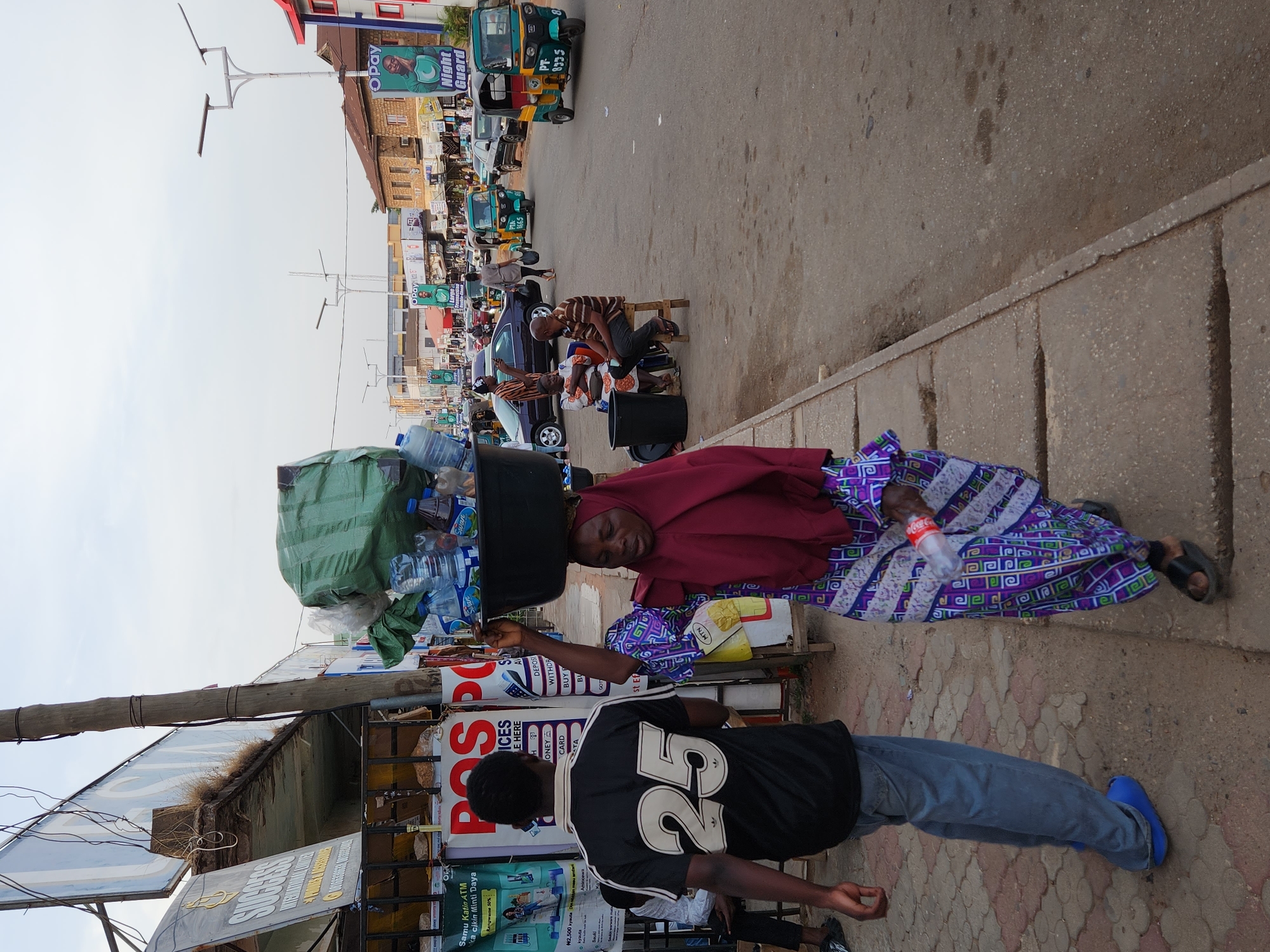 The image depicts a street scene in Jos, Nigeria. An African woman is balancing a large black tub filled with bottled water and other goods on her head. She is wearing a maroon headscarf, a patterned purple and white dress, and black sandals. She is holding a bottle of Coca-Cola in her right hand. In the foreground, a person wearing a black and white "25" jersey, jeans, and blue shoes is walking. Behind the woman, there's a POS stand and various advertisements are visible, including posters for "Success International" and "Night Guard". In the background, there are vehicles, including "keke napeps" (tricycles), other parked cars, and people sitting on the pavement. The sky is overcast, and the buildings along the street are visible.