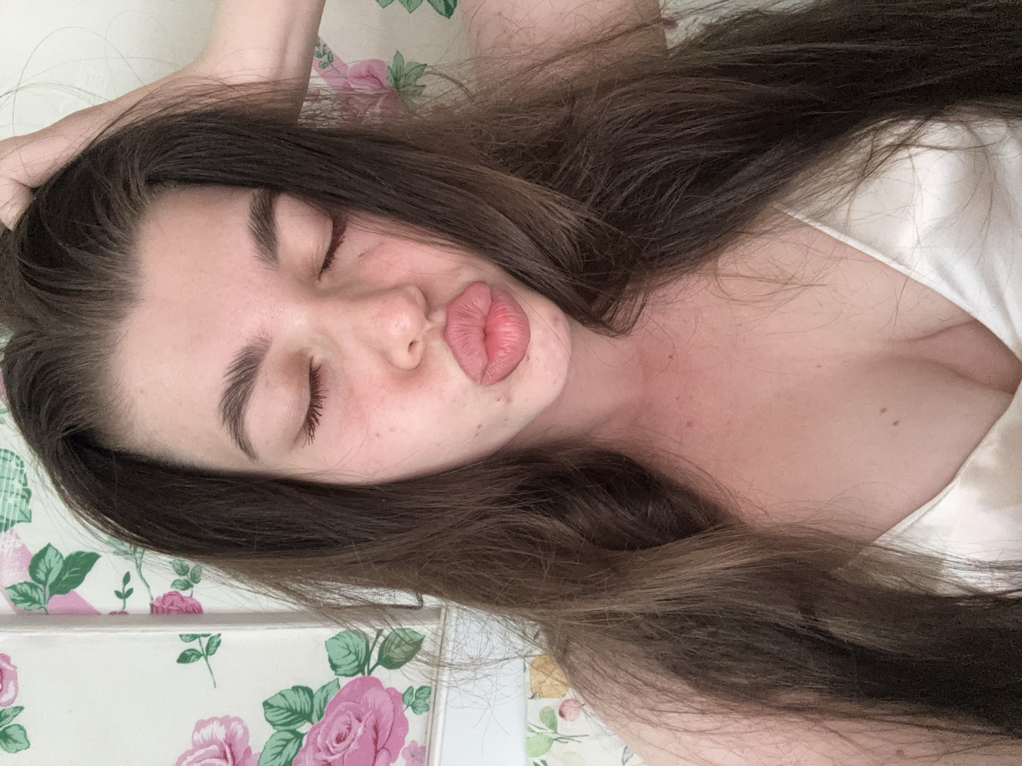 A young woman with long, dark hair is lying down. Her eyes are closed and she is pouting her lips. She is wearing a white top. The background appears to be a floral patterned surface. The location is not discernable from this image.
