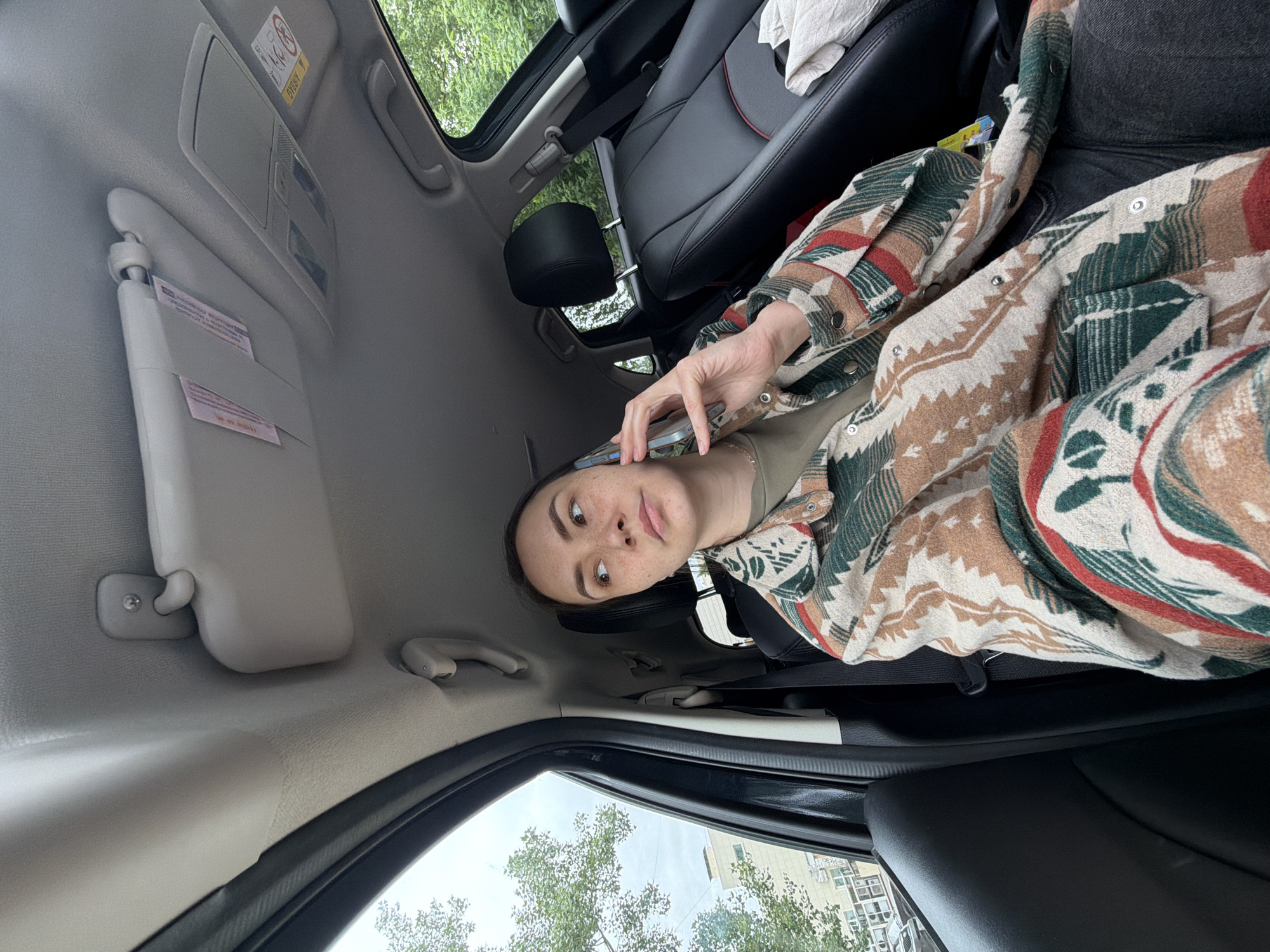 The image depicts a person seated inside a car, possibly in Chaikovsky, Russia, although there is no definitive evidence to confirm the location. The individual is holding a smartphone and appears to be talking on it. They are wearing a patterned shirt. The car's interior includes features such as leather seats, a sunroof, and a sun visor. Outside the car, trees and a building can be seen.