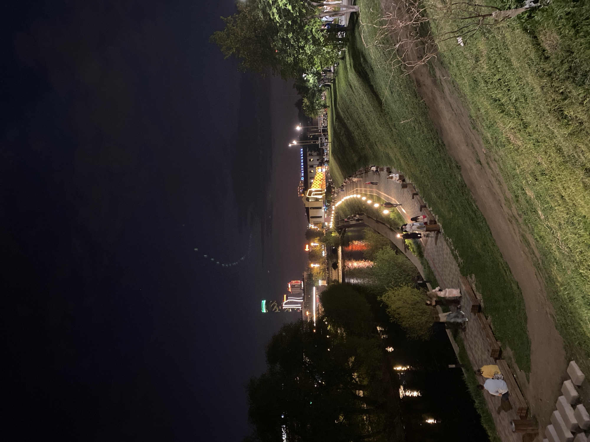 At night, an outdoor scene along the Iset River in Yekaterinburg, Russia. The left of the image is dominated by a dark night sky. From left to right, the Yekaterinburg City skyscrapers rise in the distance. A pedestrian promenade follows the Iset River's edge, with arching string lights illuminating people strolling, resting on benches, and interacting. Lush green grass slopes upwards from the promenade, extending towards trees along the upper edge. Street lights provide further illumination. The Iset River is adjacent to the walking path and reflects the lights of the city in its water.