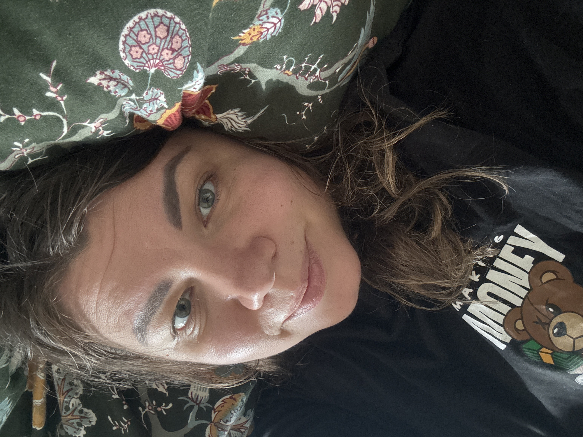 This is a close-up, portrait-oriented selfie of a woman lying down, with her head resting on a patterned pillow. The pillow features a dark green background with a repeating floral and bird motif in shades of pink, blue, red, and yellow. The woman has light brown hair, which is slightly messy and falls across her face and shoulder. Her skin appears smooth with some freckles visible. She is wearing a black t-shirt with a graphic print. The print includes the text "MAKE MONEY" in large, white, block letters. To the right of the text is a cartoon illustration of a brown bear wearing a gold chain, sitting on a pile of green money. The woman's expression is relaxed and she appears to be smiling slightly. Her eye makeup is noticeable, with defined eyebrows and eyeliner. The lighting suggests it might be daytime indoors, possibly in a bedroom or living space given the presence of the pillow. The overall mood is casual and comfortable. The location context provided is Höhenkirchen-Siegertsbrunn, Germany.