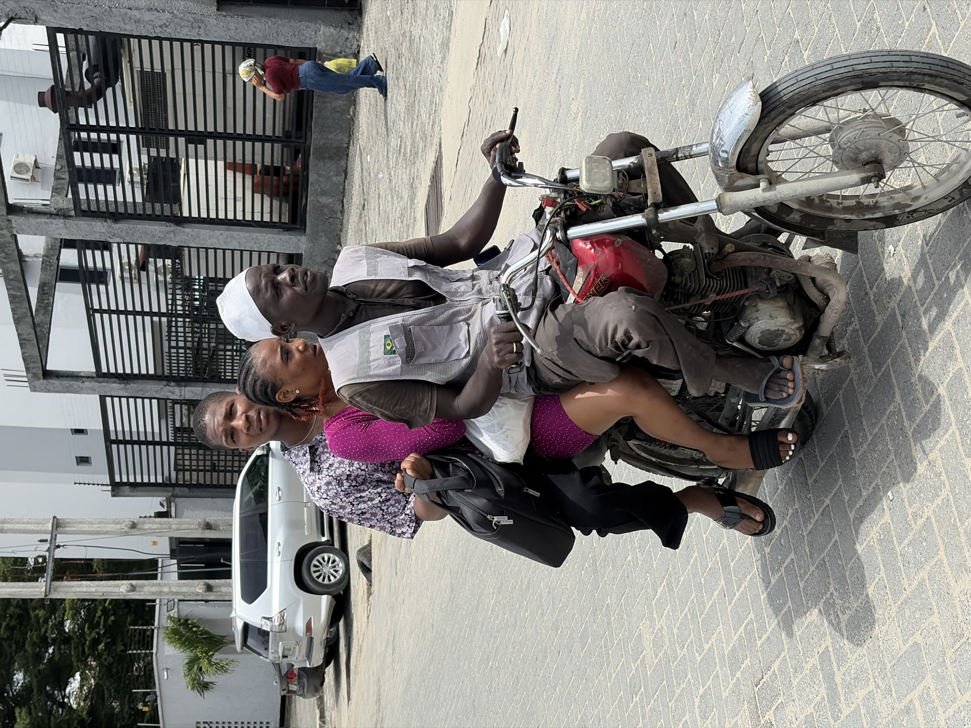 A street scene in Igboefon, Nigeria, unfolds in bright daylight under a clear sky, evidenced by the strong shadows on the herringbone-patterned paved ground. Three individuals are riding a motorcycle, while others are visible in the background.
The central figure is a man driving the motorcycle, wearing a white skullcap, a light-colored vest over a dark shirt, and dark trousers. A distinctive patch resembling the Brazilian flag is visible on his vest. He wears a ring on his right hand as he grips the handlebars of the red-tanked motorcycle, which appears to be an older model with visible engine components. Seated directly behind him is a woman wearing a vibrant magenta top with white polka dots and dark shorts or a skirt. She has visible jewelry, including an earring and a necklace. The third person on the motorcycle is another woman, positioned behind the first passenger. She wears a patterned white and dark top and carries a black shoulder bag or briefcase. All three individuals on the motorcycle are wearing sandals.
In the mid-ground, a white SUV-type car is parked. Further in the background, another person, dressed in a red shirt, blue jeans, and a light-colored headwrap, stands next to a building featuring prominent black slatted fences, holding a yellow bag. A partial figure wearing a hat is also visible behind the fence. The scene captures a moment of daily life, likely transportation, in an urban or semi-urban setting. No other explicit text is clearly legible beyond the possible "EVER" on the motorcycle's tank and the Brazilian flag emblem.