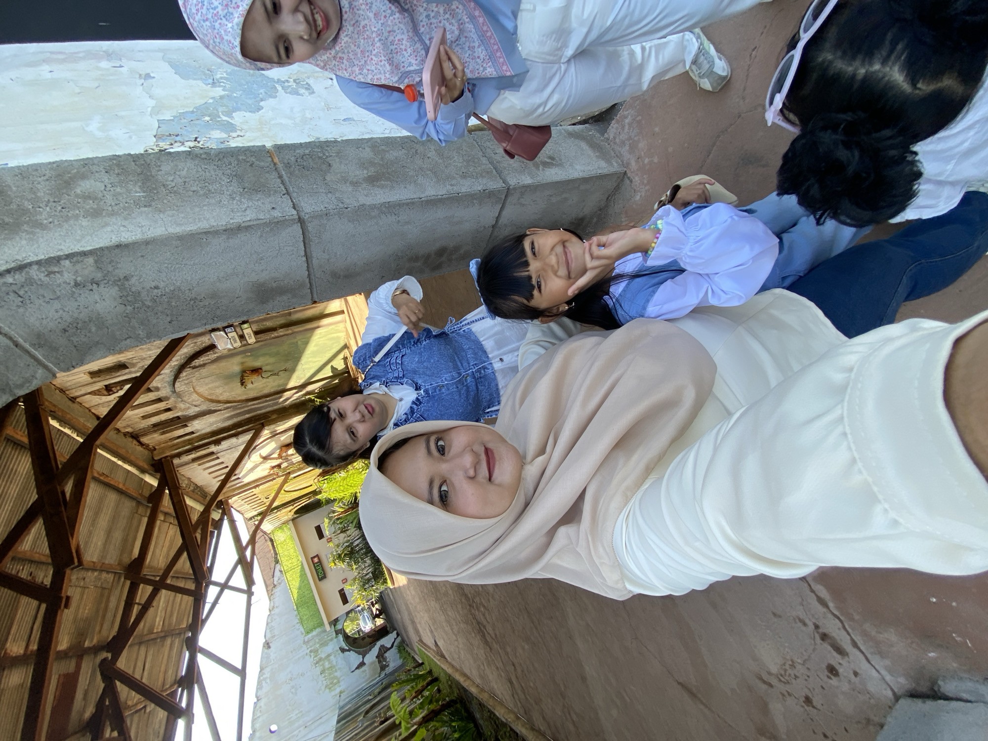 The image is an eye-level selfie of a group of four people, likely women and girls, outdoors in Mangli, Indonesia. The primary subject, presumably holding the camera, is a woman wearing a cream-colored hijab and a white long-sleeved garment. Behind her, there is a young girl in a blue denim jacket over a white top, making a V-sign with her fingers. To the left, a view reveals a skeletal wooden structure possibly belonging to an old building. In the background, there's a building with a sign that reads "TOILET" vertically in English. Another woman in a light blue outfit and a pink floral hijab is also visible. The backdrop includes glimpses of vegetation and what seems to be another structure, adding depth to the scene.