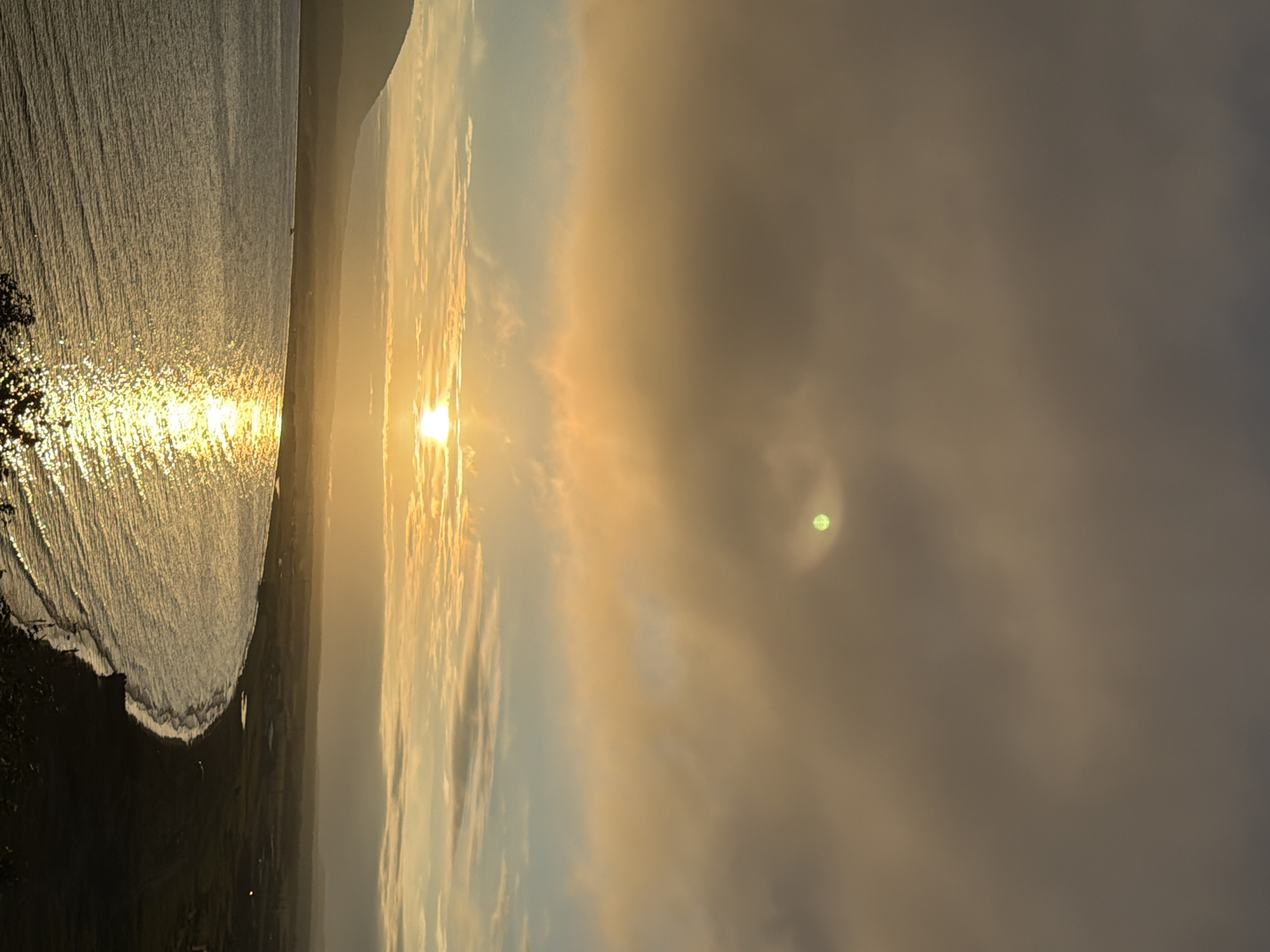 The image depicts a coastal landscape at sunset, likely in the vicinity of Yuzhno-Kurilsk, Russia, given the curvature of the land and the presence of a body of water that appears to be the Sea of Okhotsk. The sun, partially obscured by clouds, casts a brilliant golden light across the sky and reflects intensely on the rippling surface of the sea. A mountainous silhouette is visible on the left horizon. The coastline features a dark beach with sparse vegetation and a few distant, low-lying structures suggesting a settlement or industrial area. The sky is filled with layers of clouds, ranging from a deep, muted gray at the top to lighter, warmer tones of orange and yellow closer to the horizon, with a faint greenish circular anomaly in the upper sky. The overall scene conveys a sense of natural beauty and vastness.