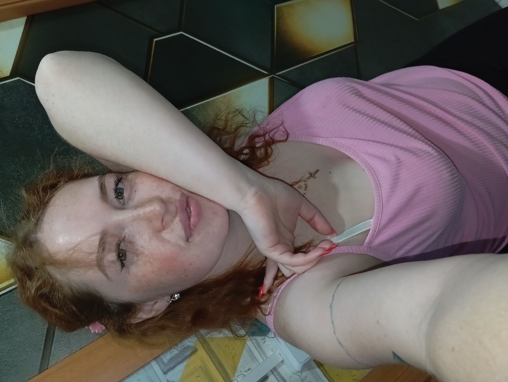 A woman with red hair and freckles is lying on a surface with a geometric tile pattern. She is wearing a pink top and has her arm raised, with her hand near her face. The background includes a dark area, possibly a wall or furniture. The location is unknown.