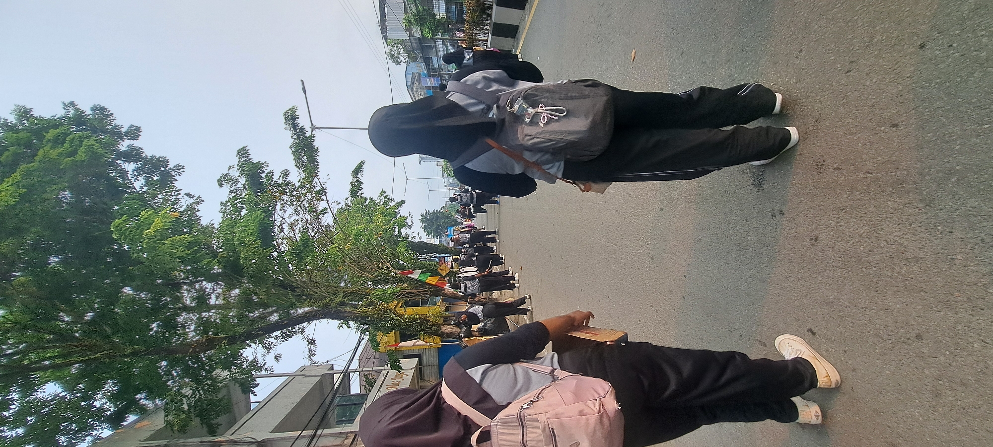Two young women, both wearing hijabs and athletic attire, are walking on a paved road in Tarakan, Indonesia. The woman on the left has a pink backpack and is holding a flat, rectangular object. The woman on the right has a dark gray backpack. They are walking away from the camera, on the side of a road with trees lining the left side and buildings and a street lamp visible in the background. Further down the road, a marching band or drum corps can be seen in formation, with individuals holding instruments such as drums. The sky is overcast and hazy. The environment appears to be an urban setting.