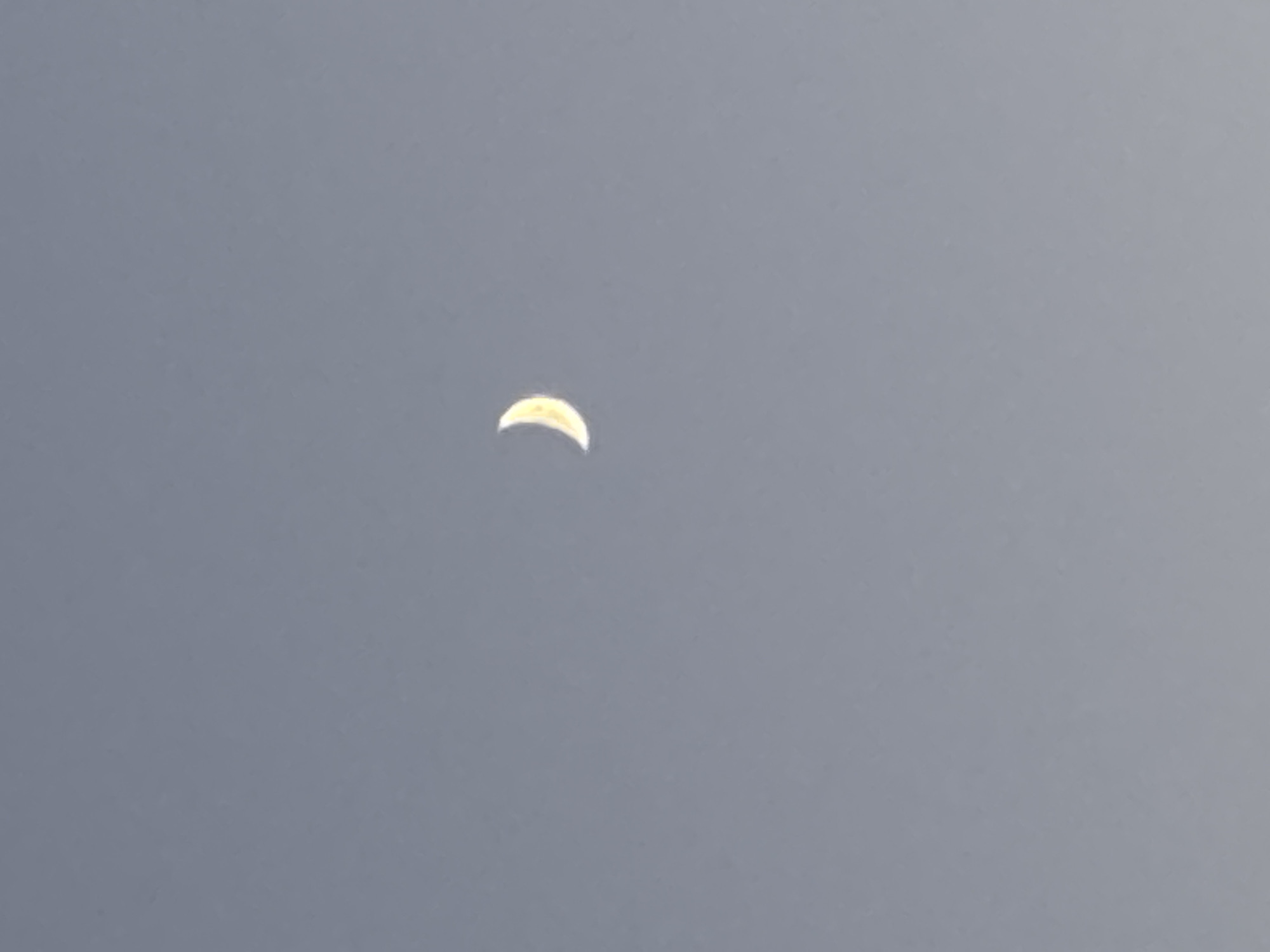 The image is a photograph of the moon in a clear, pale grey sky. The moon is a crescent shape, with the illuminated portion being a bright yellow-white. The moon is positioned slightly off-center to the right and in the upper half of the frame. There are no other discernible objects or features in the sky. The setting appears to be outdoors during twilight or a hazy daytime. No specific location in Minsk, Belarus, can be identified from this image.