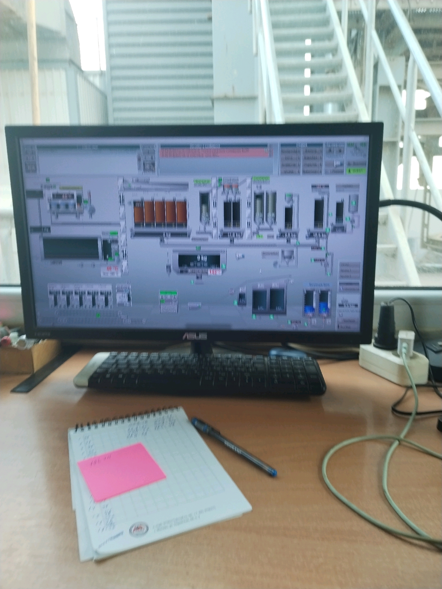 The image displays a desktop computer monitor showing a complex industrial control interface, likely for a chemical plant or manufacturing facility. The screen features various graphical elements representing processes, equipment, and numerical data, such as tanks, pipes, and flow meters. To the right of the monitor, a white power strip with two plugs and a black adapter is visible, connected to power cables. In front of the monitor sits a black computer keyboard. To the left of the keyboard, a spiral-bound notebook is open, with handwriting on its pages and a pink sticky note placed on top. A blue pen rests on the notebook. The scene is set on a wooden desk. In the background, out of focus, appears to be an industrial setting with metal structures, possibly a staircase and siding, suggesting an indoor industrial environment. There are no people visible in the image. The overall impression is that of an operational control room or a workstation within an industrial complex.