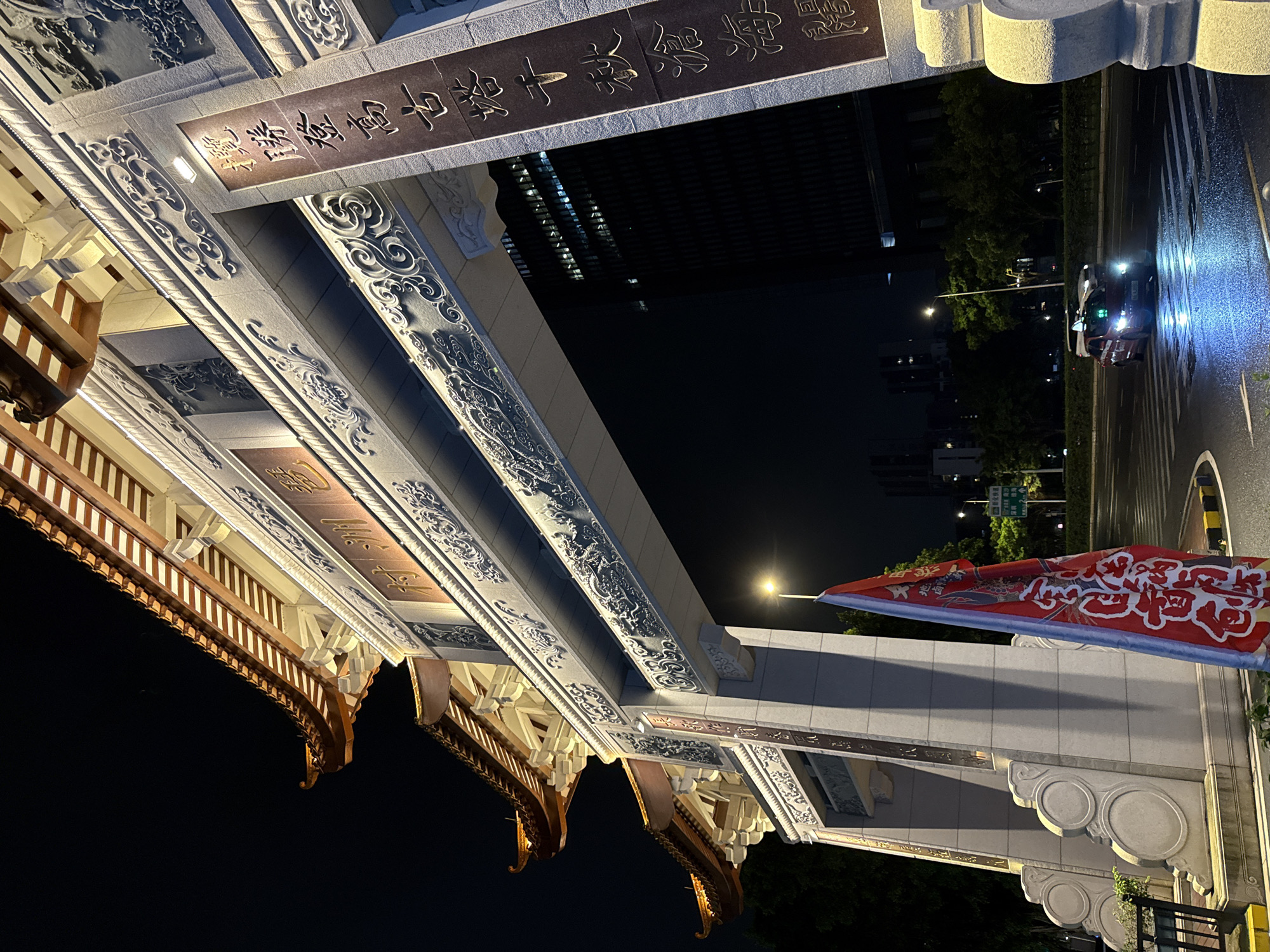 An ornate, illuminated archway in the Haizhu District of China stands at night. The archway features traditional East Asian architectural elements, including tiered, upturned eaves with reddish-brown tiles and intricate decorative carvings along its facade. Chinese characters are prominently displayed on vertical plaques on either side of the arch. To the right of the arch, a red banner with white Chinese calligraphy is affixed to a pole, slightly obscuring the view of the arch. Beyond the arch, a modern, dark skyscraper with illuminated windows is visible. A street in front of the archway is wet, reflecting the lights of a red vehicle, likely a taxi, crossing the street. Trees line the background.