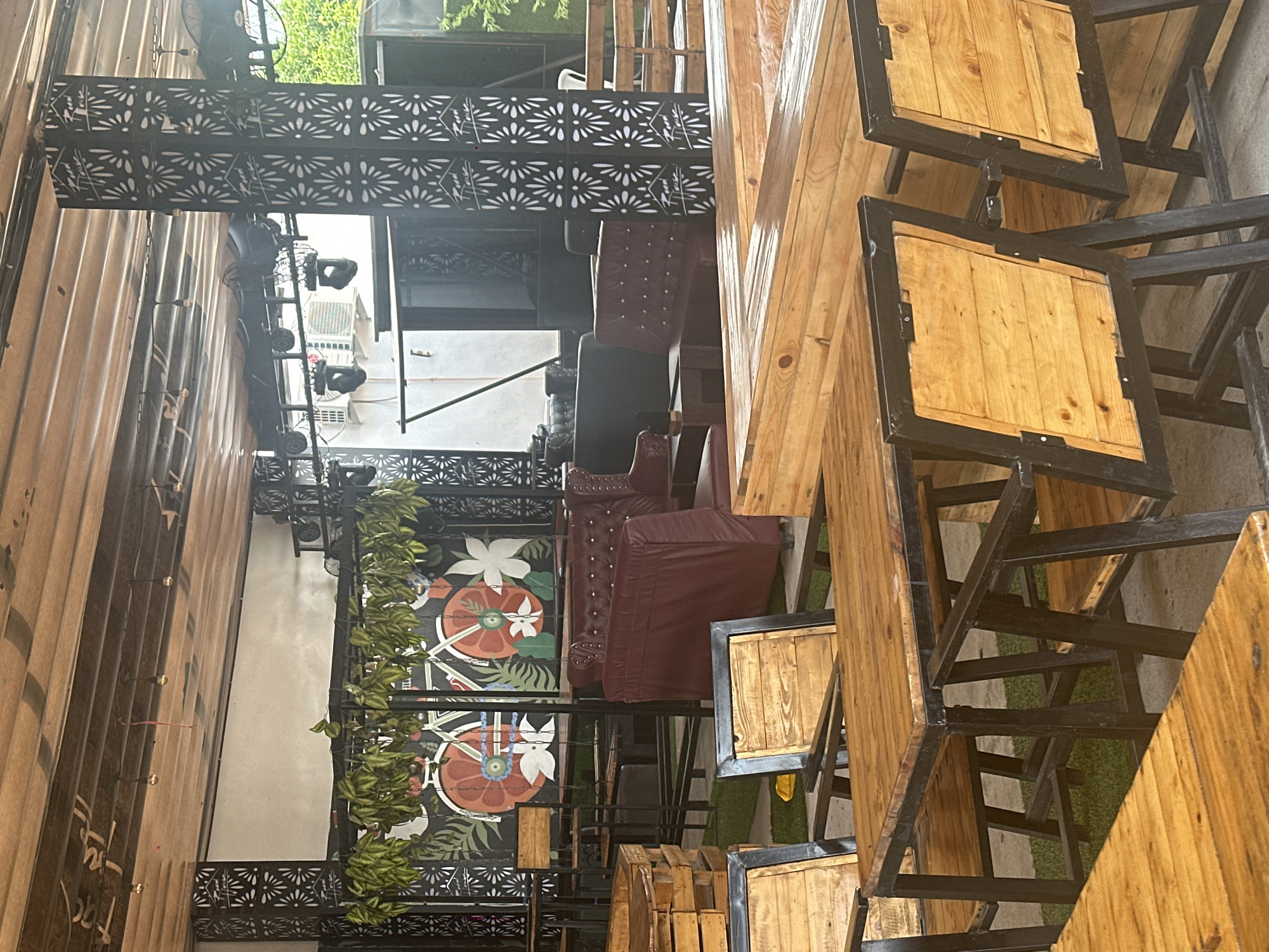 The image depicts an outdoor seating area, likely a cafe or restaurant, with a rustic industrial design. The furniture is predominantly made of wood and metal, featuring sturdy tables and chairs. In the background, there are plush, burgundy leather sofas and armchairs arranged in a lounge setting. Decorative elements include hanging plants and a mural featuring a stylized bicycle and floral motifs. The ceiling is constructed of wooden planks with exposed beams and lighting fixtures. The space is defined by black metal pillars with intricate cut-out patterns. The overall ambiance suggests a trendy and casual dining or social gathering spot. The location is Howayi, Nigeria. There are no people visible in the image.
