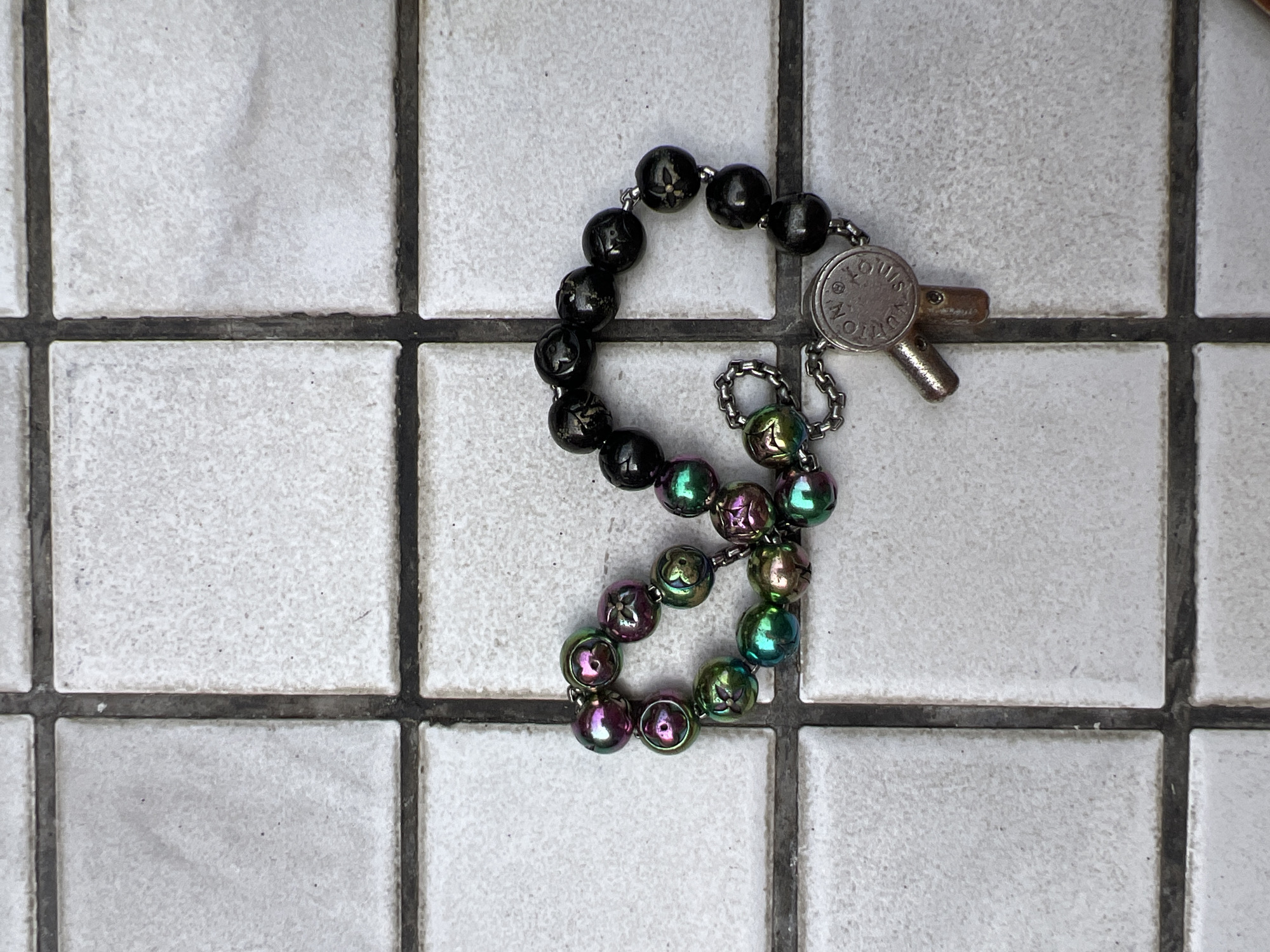 A bracelet with black and iridescent beads is displayed on a tiled surface in Moscow, Russia. The bracelet features a small metal charm resembling a key with an embossed circular logo. The tiles are a pale gray with dark grout lines. The lighting suggests an outdoor or well-lit indoor environment.