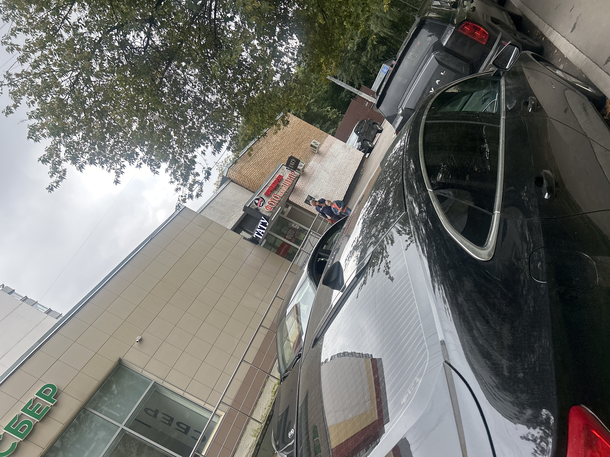 The image depicts a street scene in Moscow, Russia, with a focus on several parked cars. In the foreground, the rear portion of a black sedan is visible, reflecting the surrounding environment on its shiny surface. To its right, a black SUV is parked, with the word "VAL" visible on its rear. Further down the street, the facade of a commercial building is seen. A green sign with the Cyrillic letters "СБЕР" is prominently displayed on the building. Adjacent to it, another building displays signs for a "TATU" tattoo parlor and a "ФОТОКОПИЦЕНТР" (photocopy center). Two individuals, an adult and a child, are standing near the entrance of the commercial establishment. The sky is overcast, and trees with green foliage line the street, indicating an outdoor urban setting.