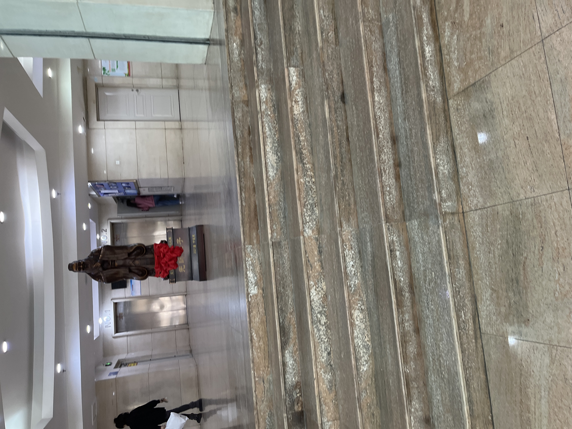 The image shows the interior of a building in Changchun City, China. The scene includes a statue of a man on a pedestal draped with a red fabric. Two elevator doors are visible behind the statue. A person is walking near the statue, and another person is standing in the distance near what appears to be a television screen. The foreground shows a tiled floor.