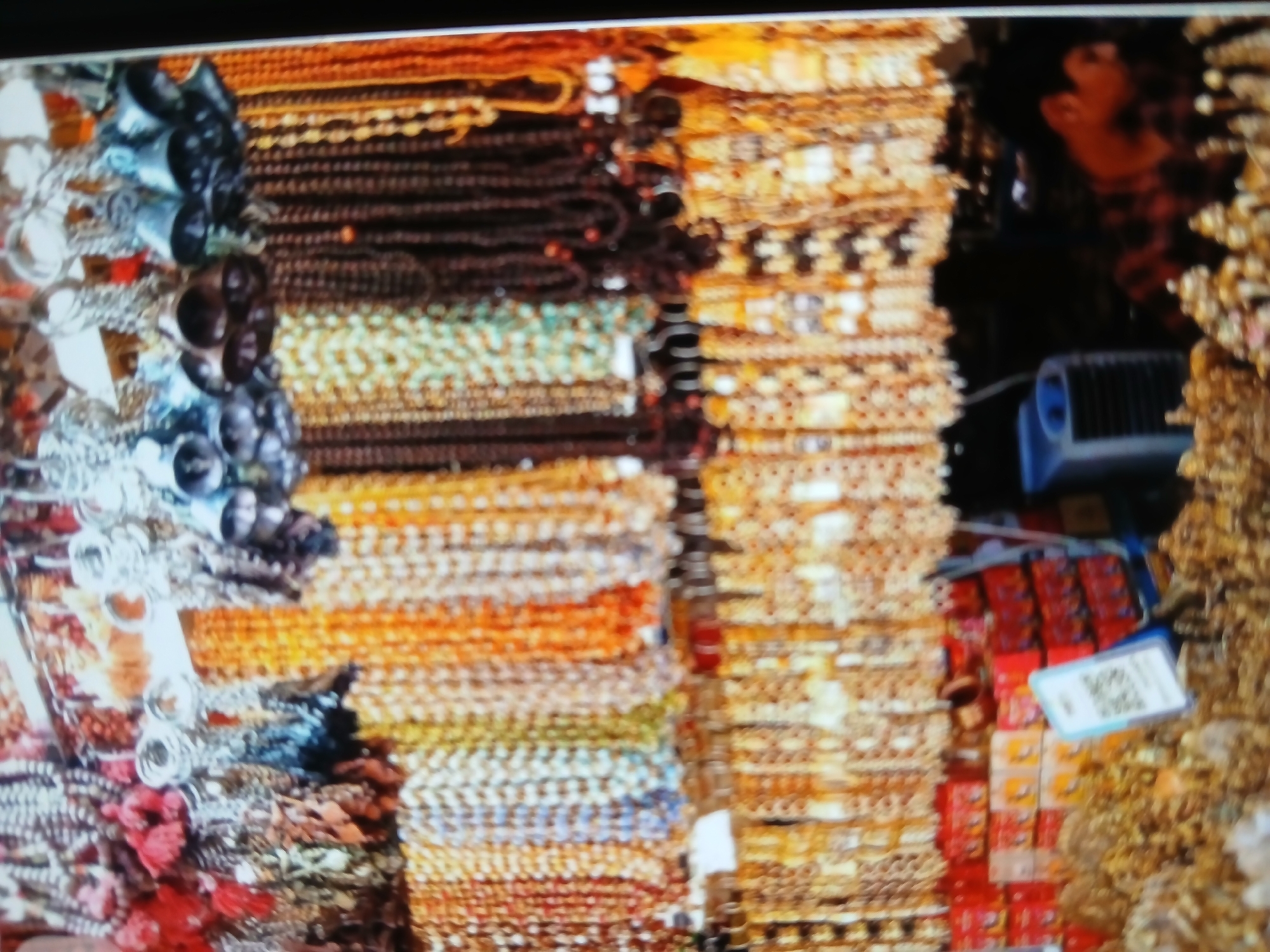 The image depicts a close-up of a retail display featuring rows of beaded necklaces and bracelets. The jewelry is arranged vertically, with various colors and designs visible. On the left side, there are items with silver and blue accents. Toward the center, the strands are mainly composed of brown, green, orange, and light-colored beads. The right side displays strands predominantly in shades of gold and brown.
In the background, there's a glimpse of a person's profile, a blue object, and small rectangular packages stacked on a shelf, with a price tag or QR code attached.
Based on the products, it can be speculated that this display is located in an outdoor bazaar, potentially in Bengaluru, India.